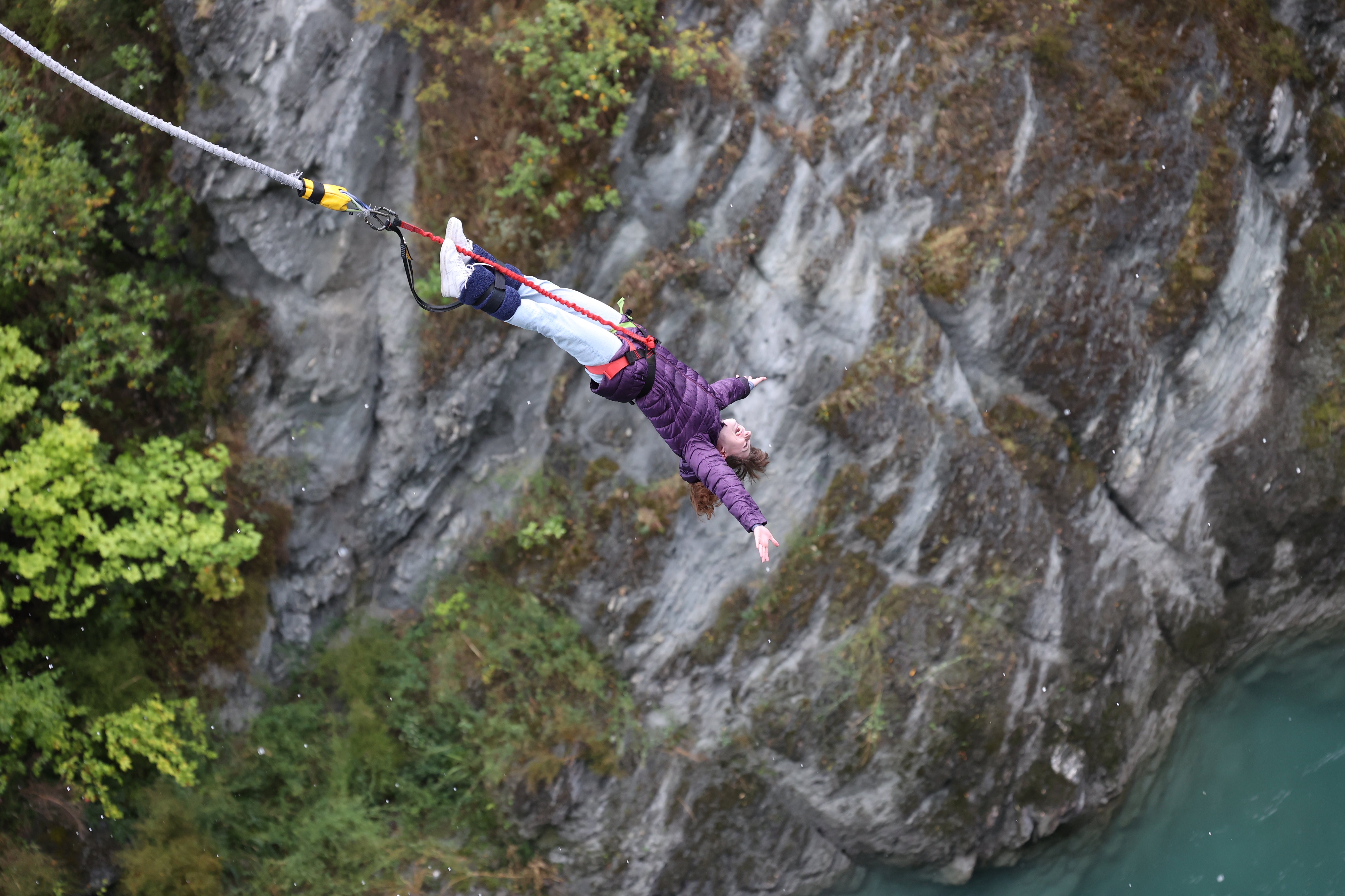 A bungee jumper wearing a purple coat and light jeans with her arms wide as she free-falls towards the Kawarau River with her bungee cords at her feet.