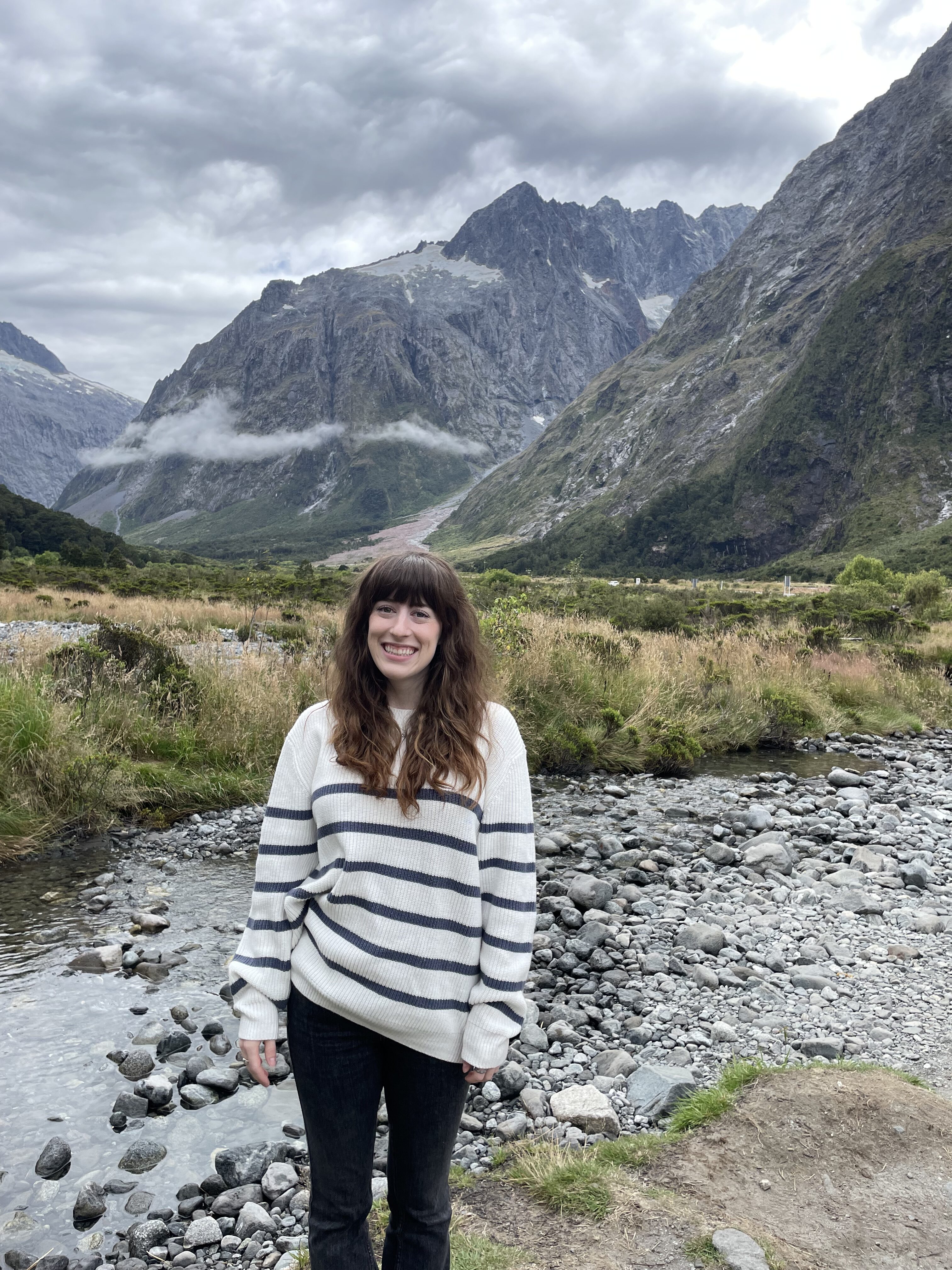 A scenic valley on the way to Milford Sound with a young woman wearing a striped blue and white sweater smiling in front of it.