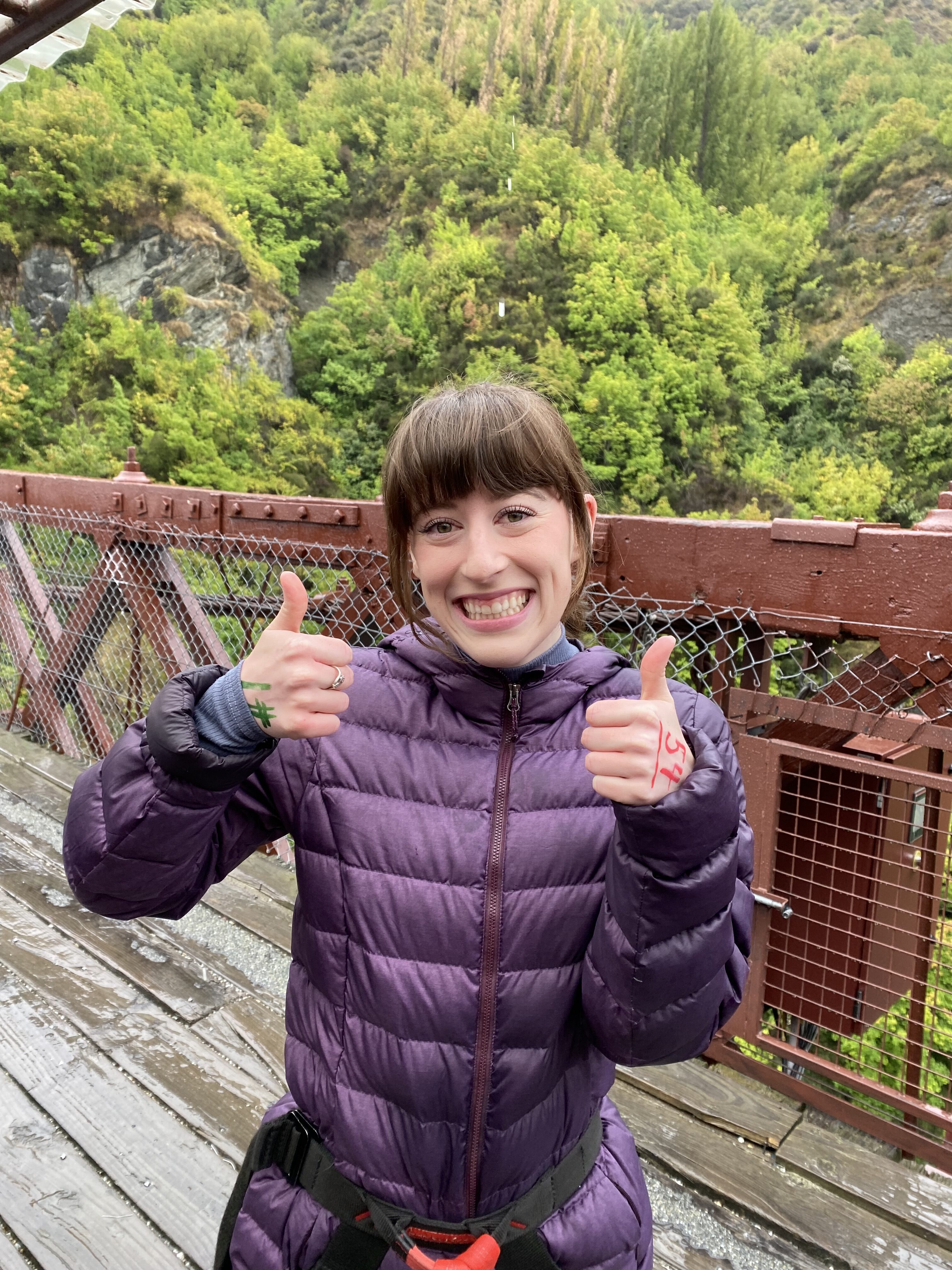 A brunette young woman with bangs wearing a purple coat giving two thumbs up at the Kawarau Bridge.