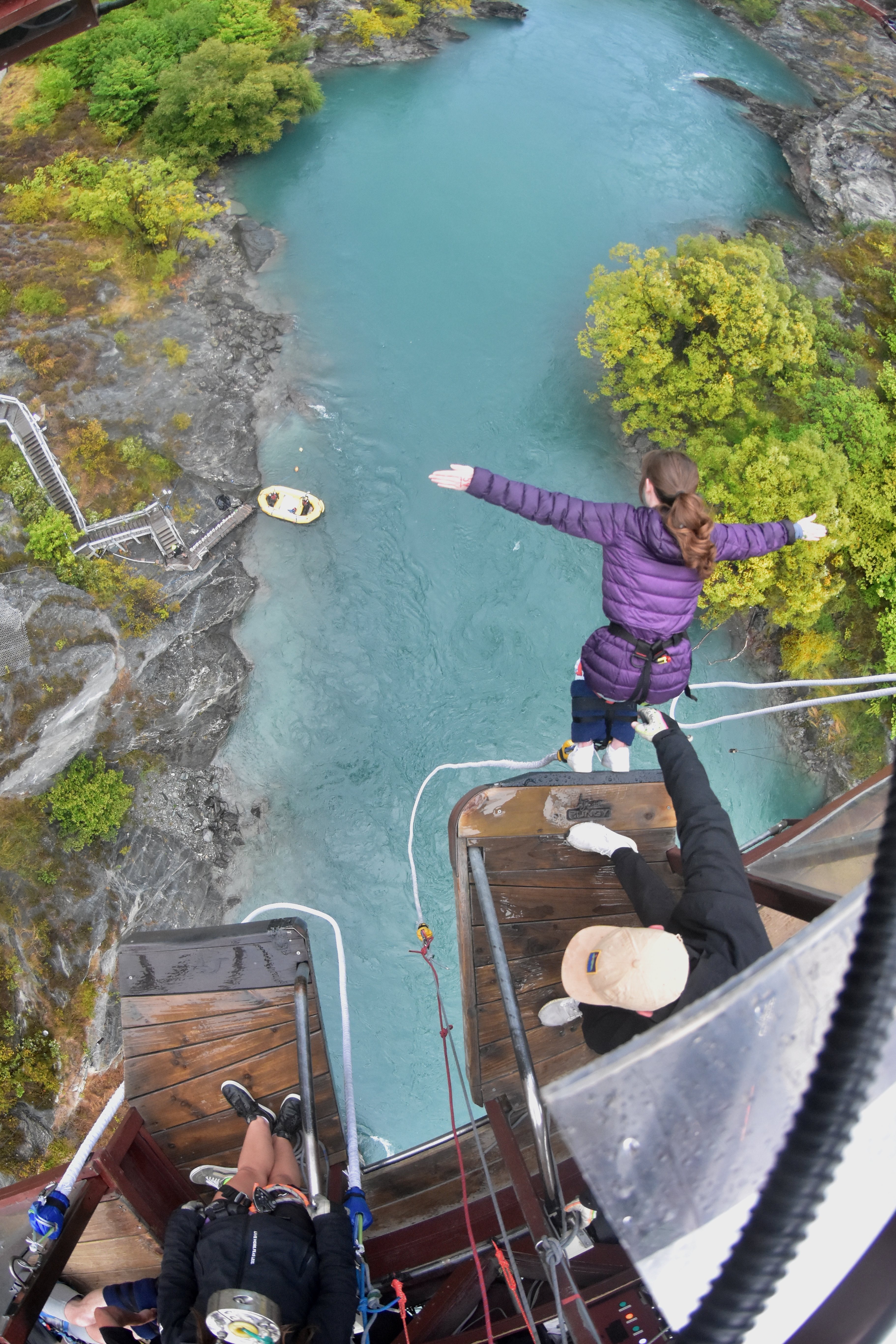 A young woman wearing a purple coat tipping off the bungee platform at Kawarau Bridge.