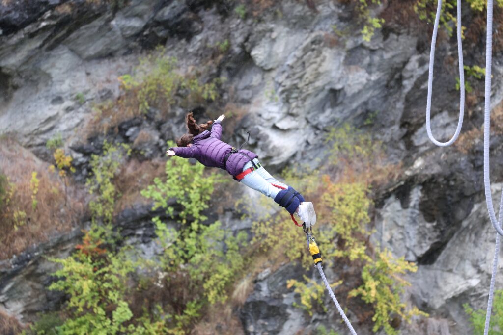 A young woman wearing a purple coat in a free fall with bungee cords tied to her feet and the rocky, shrub-covered landscape behind her.