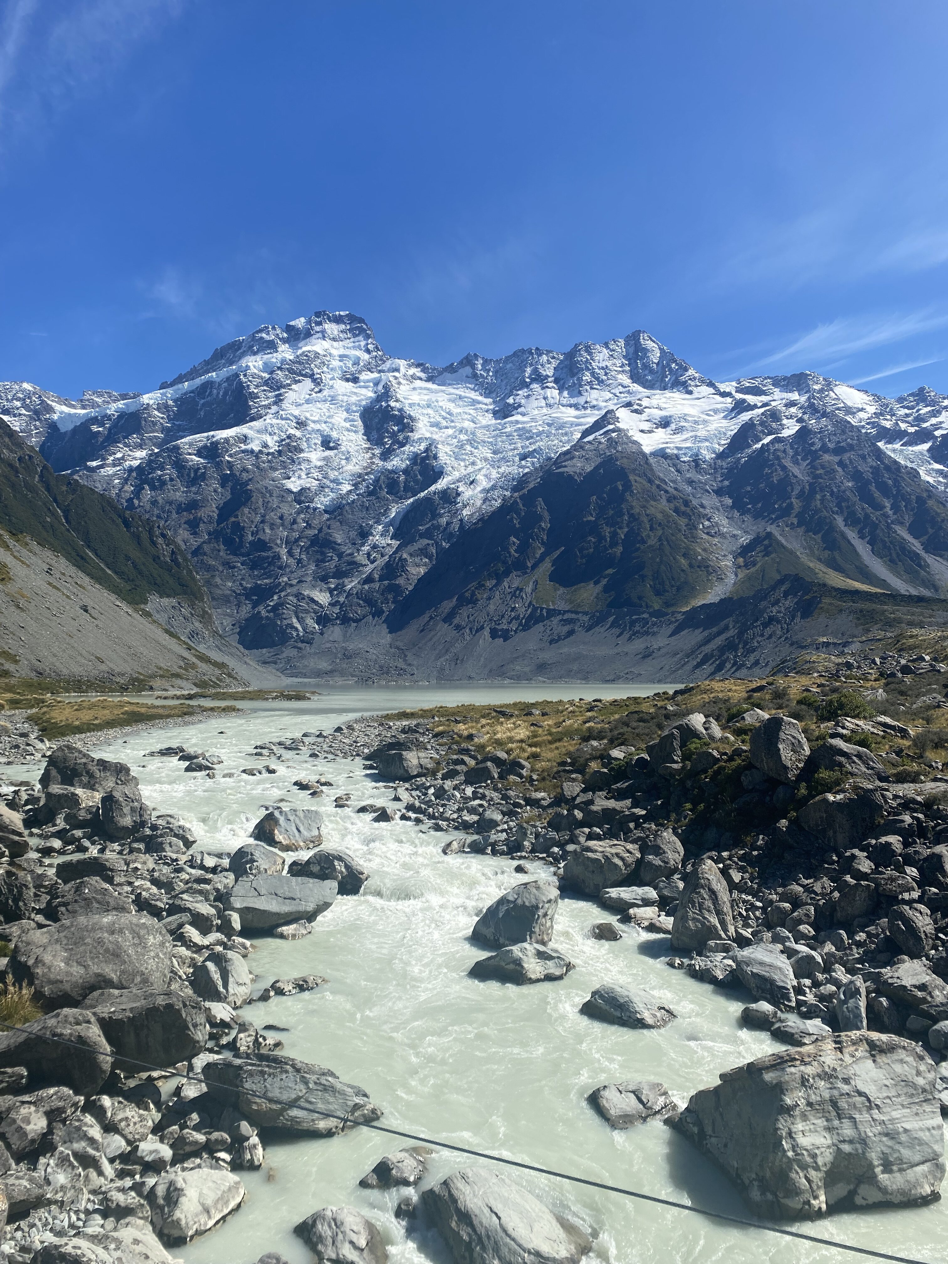 A beautiful scenic view along the Hooker Valley Track is Aoraki/Mt. Cook National Park. There is a scenic white-grey river flowing from a stunning mountain glacier.