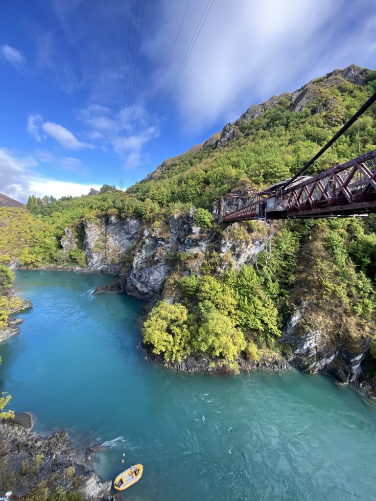 The Kawarau Bridge standing tall over the Kawarau River with a yellow raft at the rocky base.
