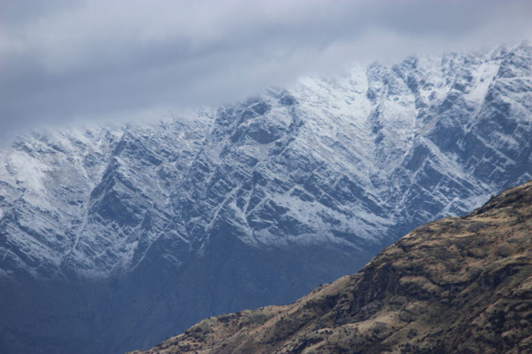 The snow-capped Remarkables framed by a tan mountainside and a grey cloud.