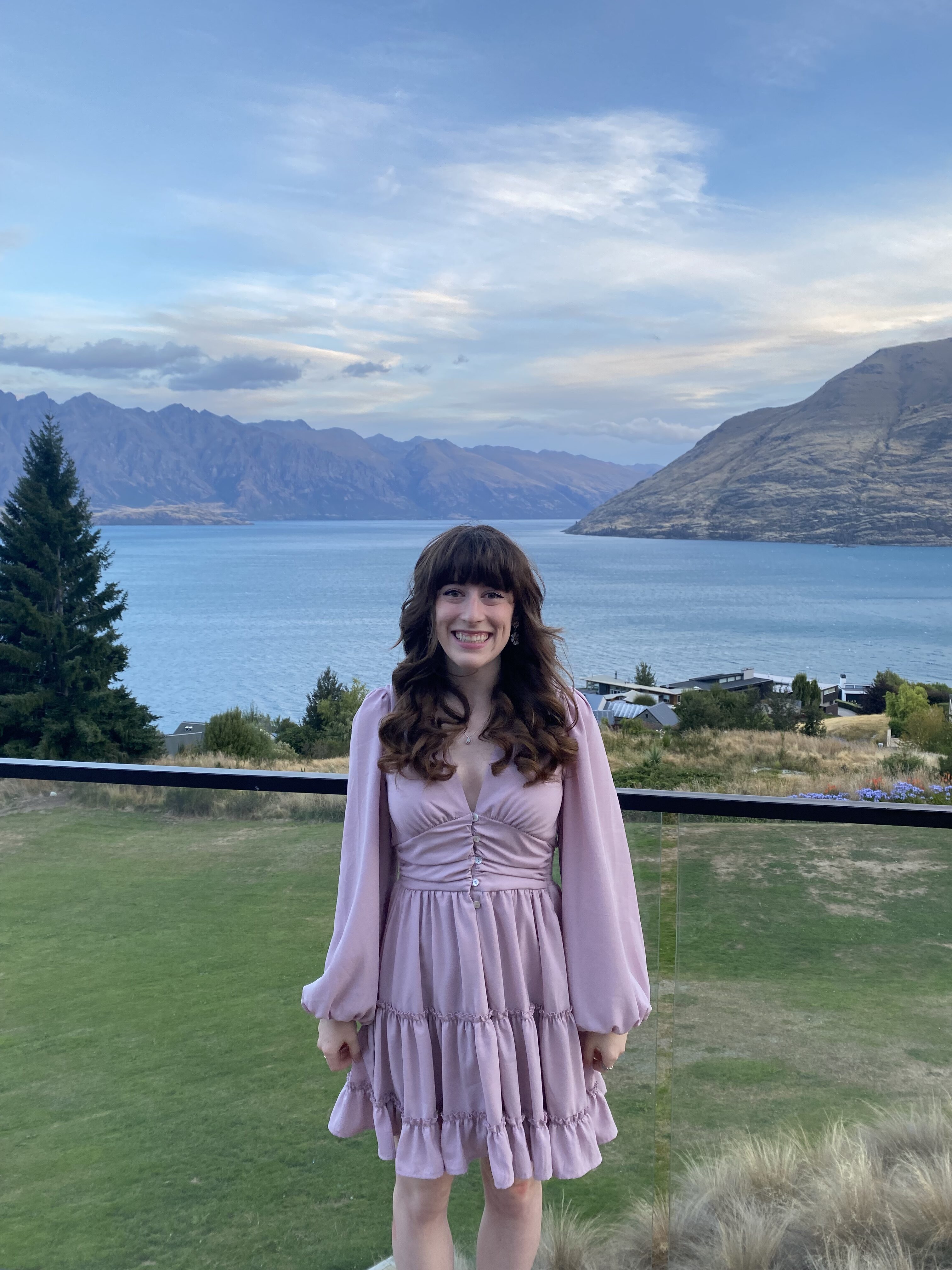 A cute brunette woman with bangs wearing a light pink dress, standing on a balcony overlooking Lake Wakatipu.