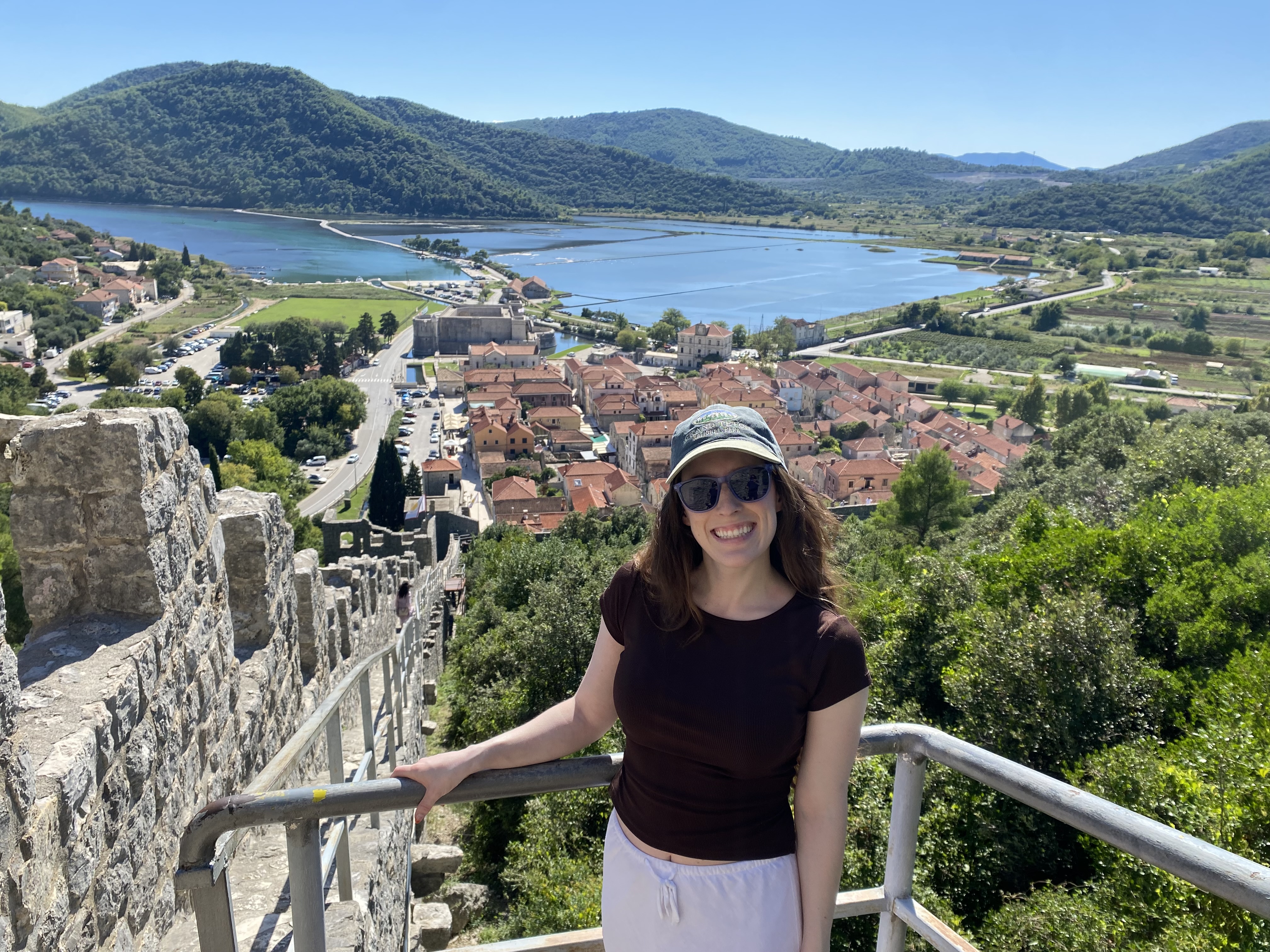 A young woman wearing a brown shirt, a ball cap and sunglasses smiling as she holds onto a silver railing on the Ston Walls, with a view of the village and the salt pans in the background.