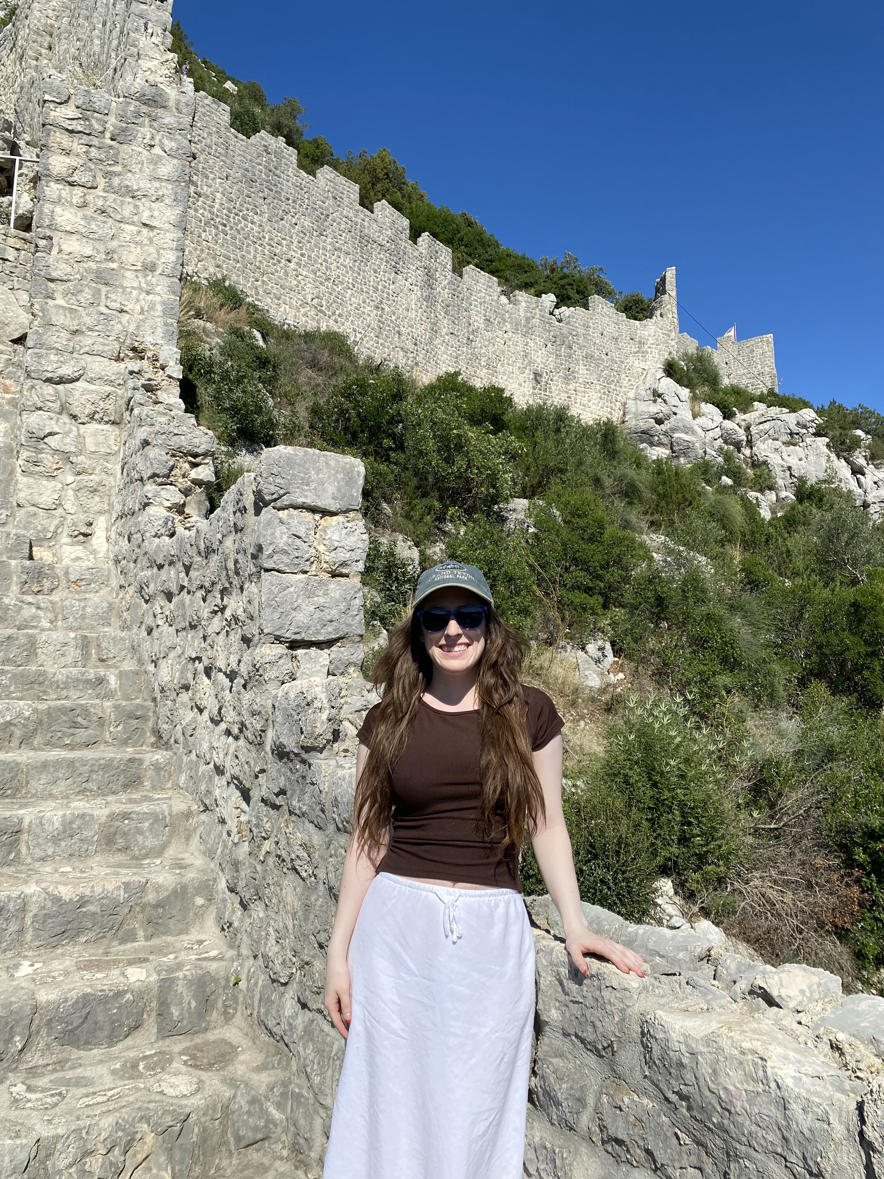 A young woman wearing a brown shirt, a white skirt and a ball cap/sunglasses smiles on the Ston Wall.