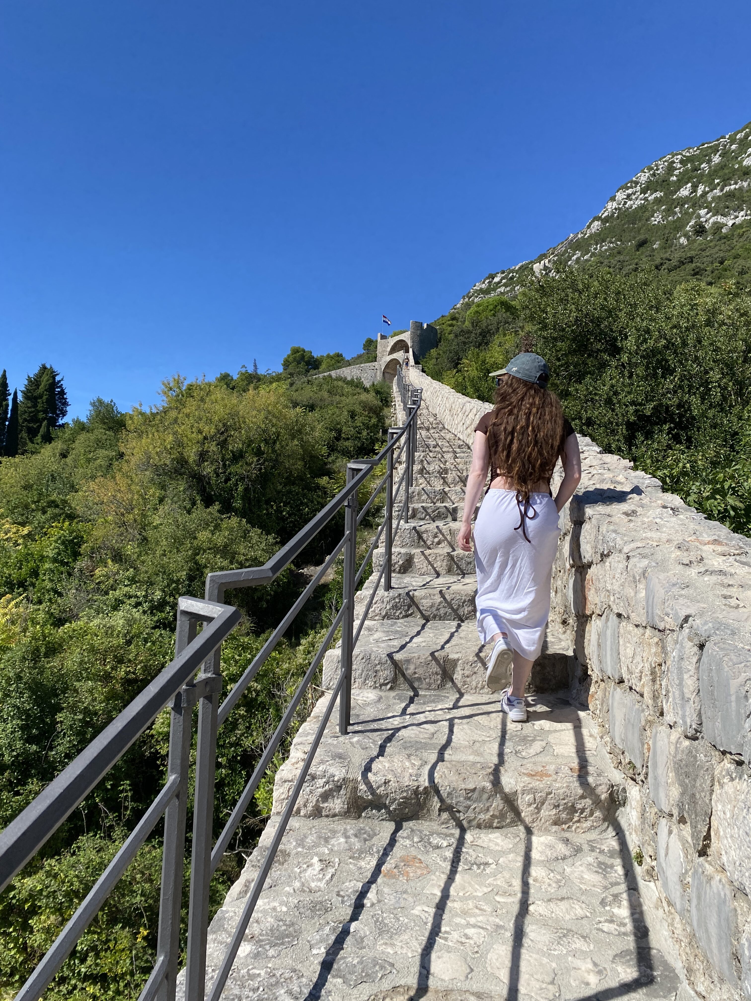 A young woman with long brunette hair wearing a long white skirt walking up the Ston Walls.