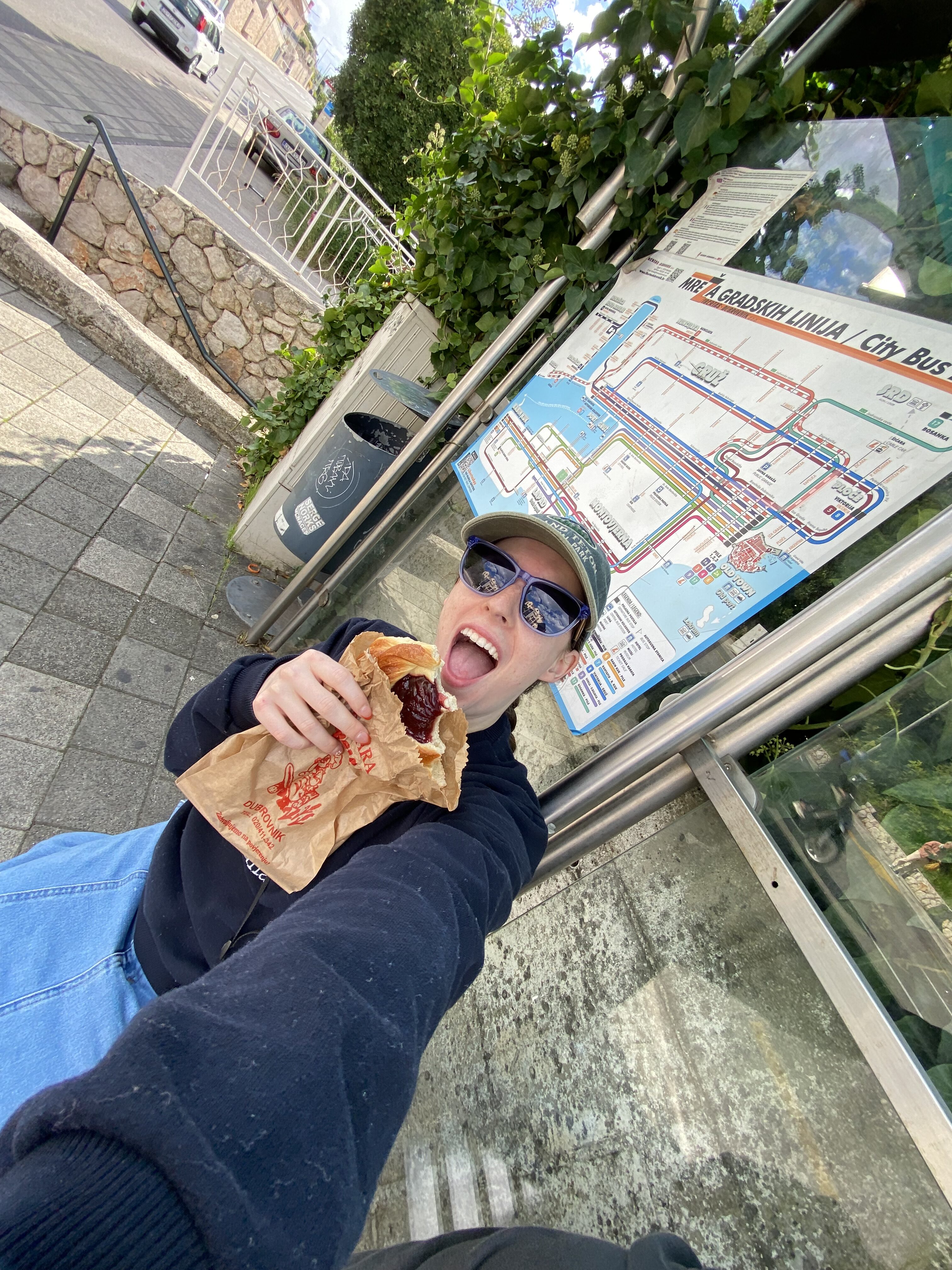 A young woman wearing sunglasses holding a pastry as she waits at the Dubrovnik bus stop.