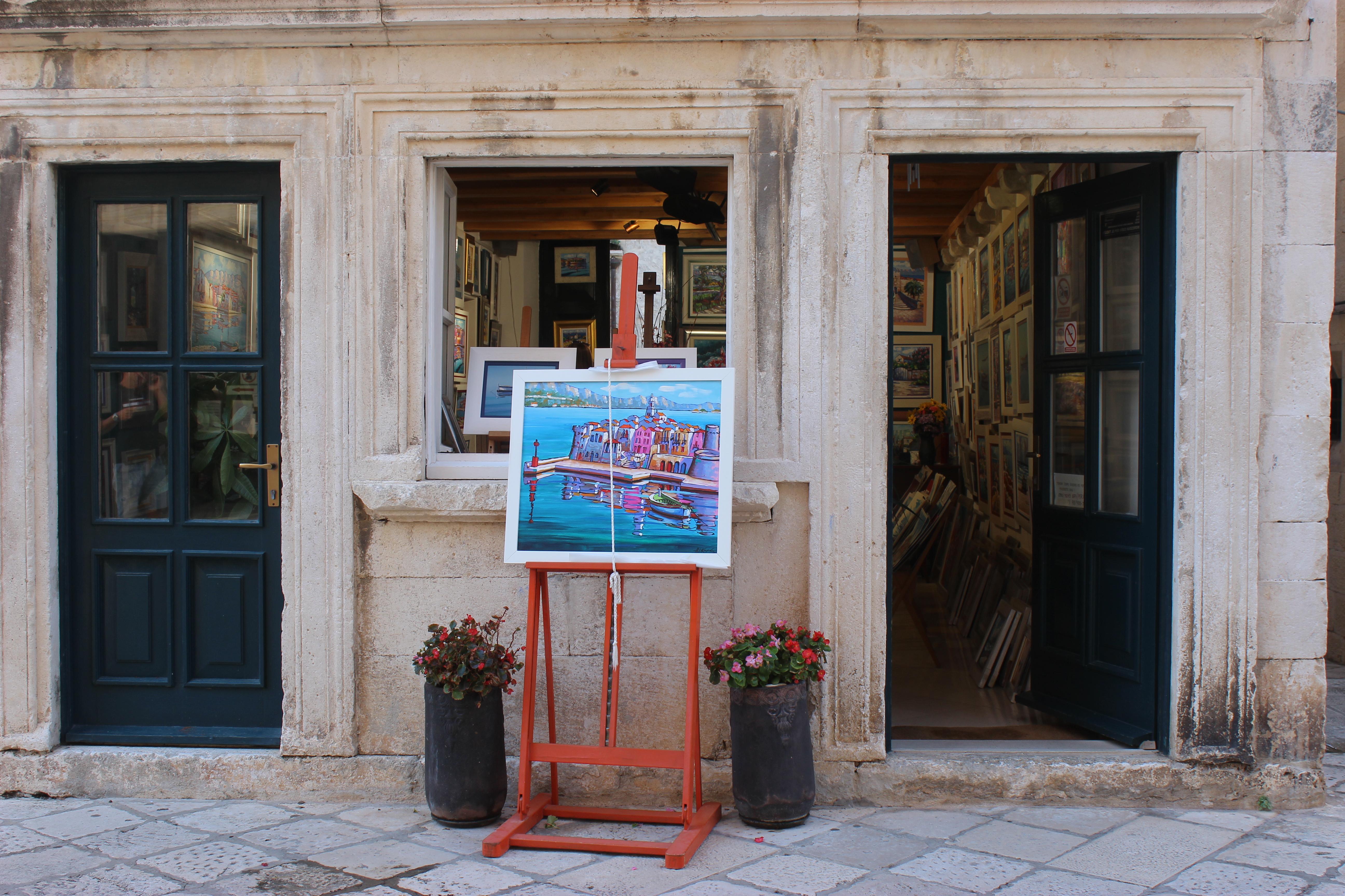 A stone art gallery in Korčula Old Town with a red easel displaying a colorful painting of Korčula Old Town.