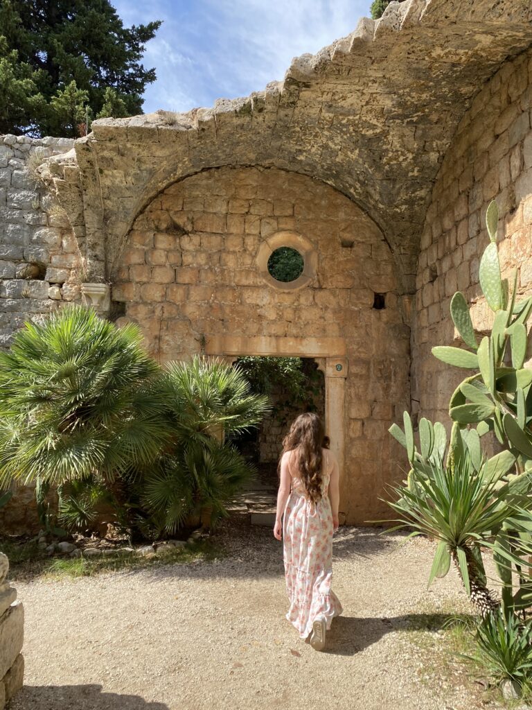 A brunette woman walking away from the camera through a dilapidated stone structure with greenery surrounding her in the monastery on Lokrum Island.
