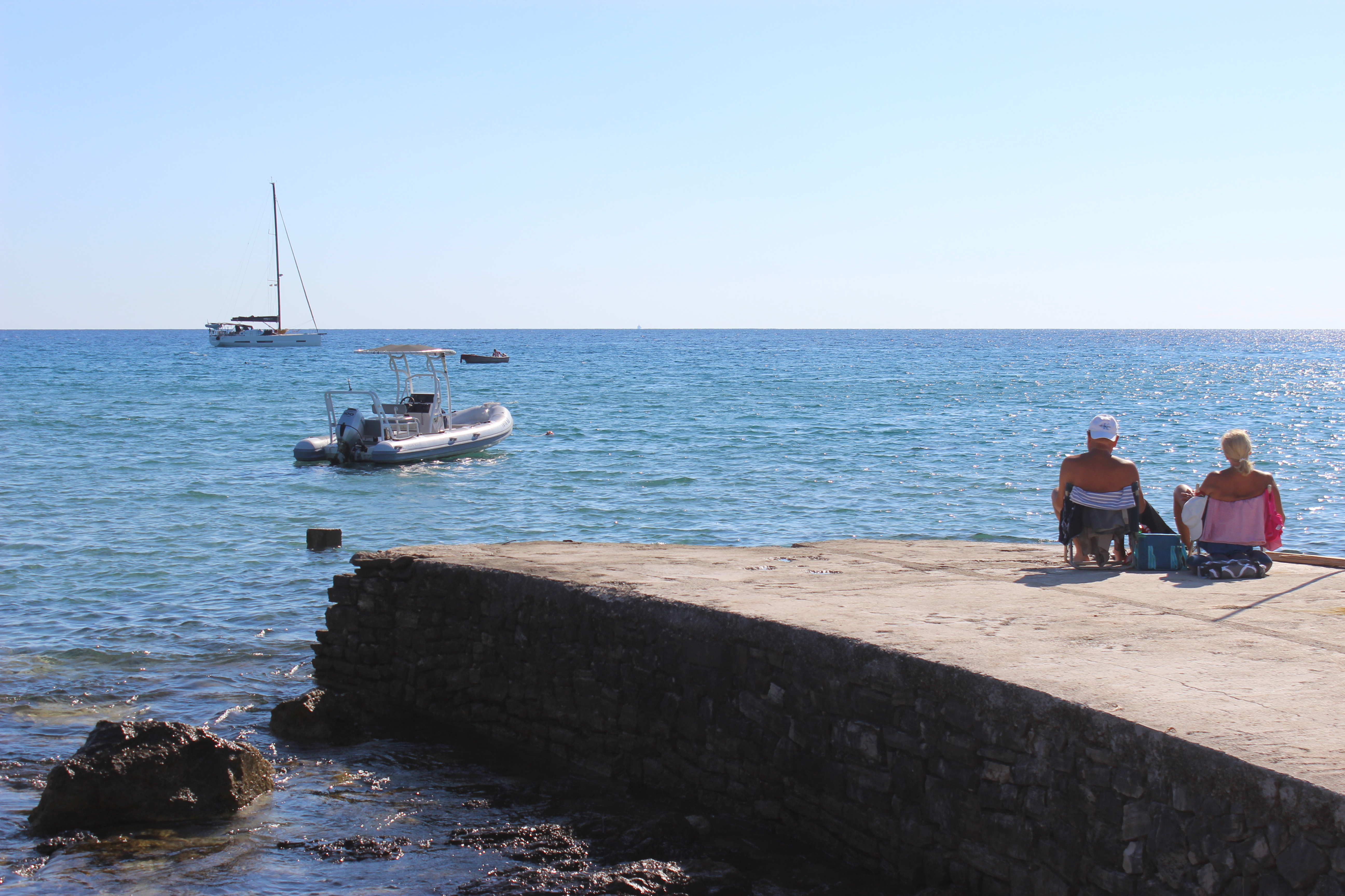 An old couple lounging on a stone dock in Korčula looking out to a small boat and a sailboat in the background.