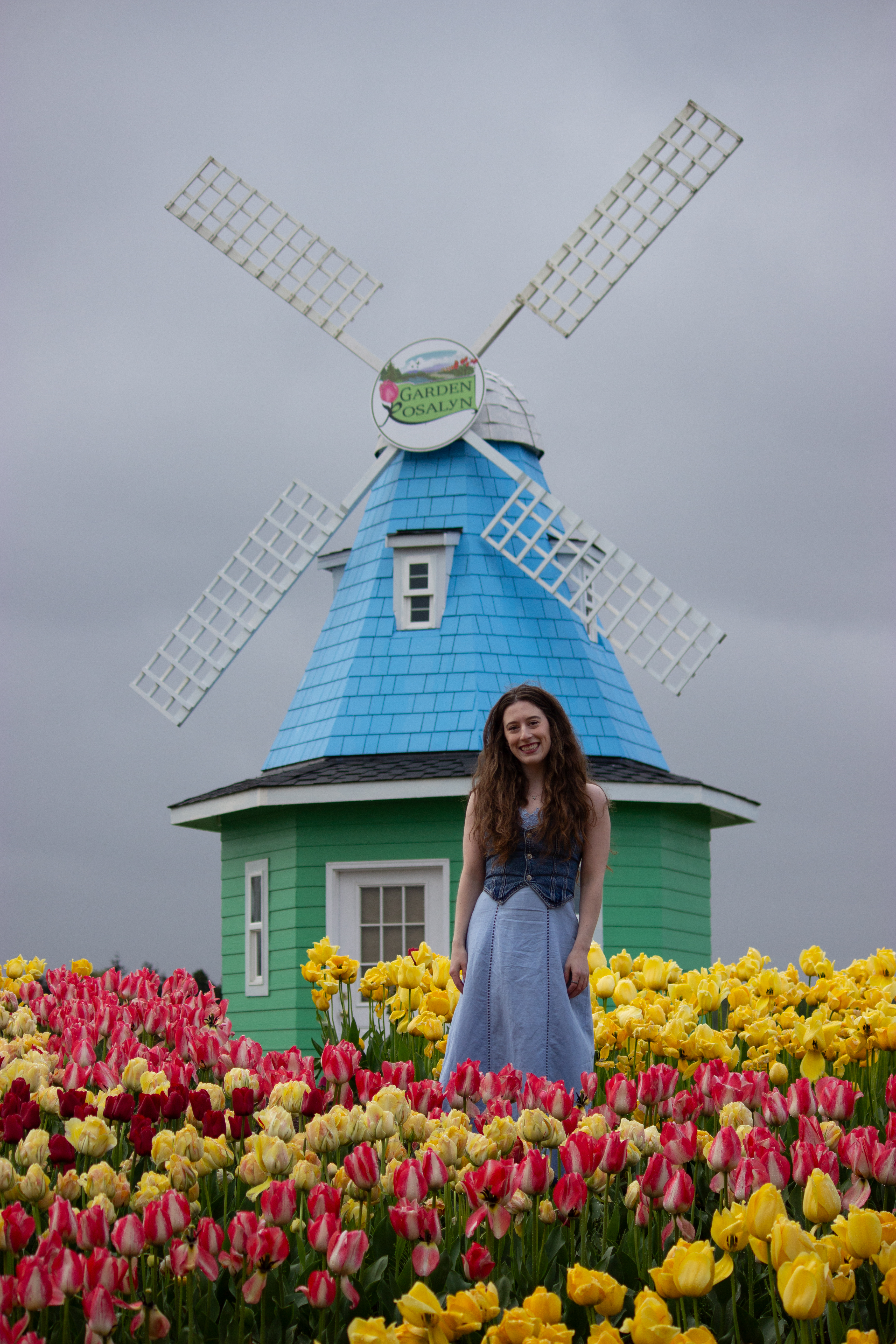 A girl wearing a blue dress and jean vest standing in front of blue and green windmill at Garden Rosalyn at the Skagit Valley Tulip Festival.