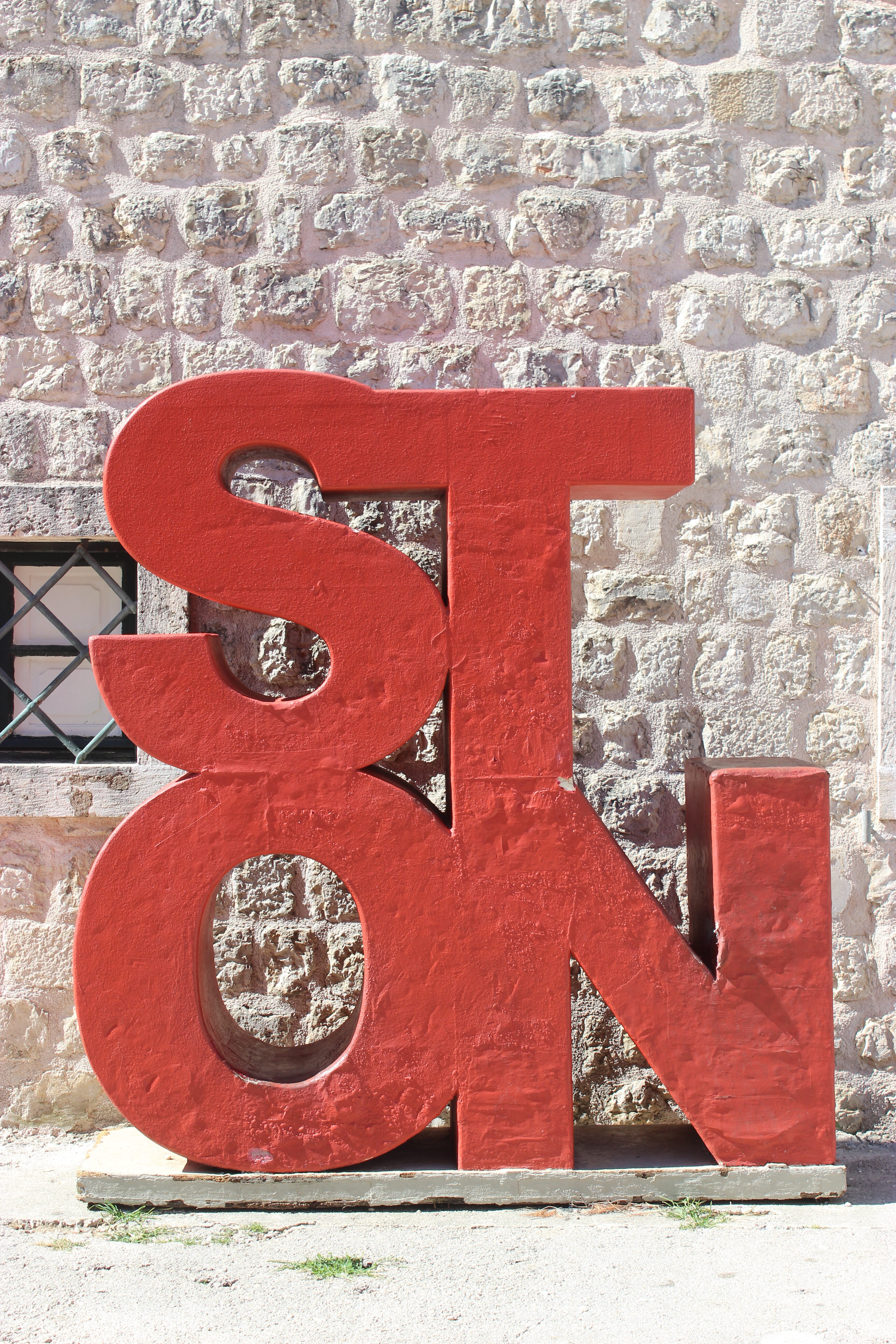A red sign that says "Ston" in front of a light grey stone wall.