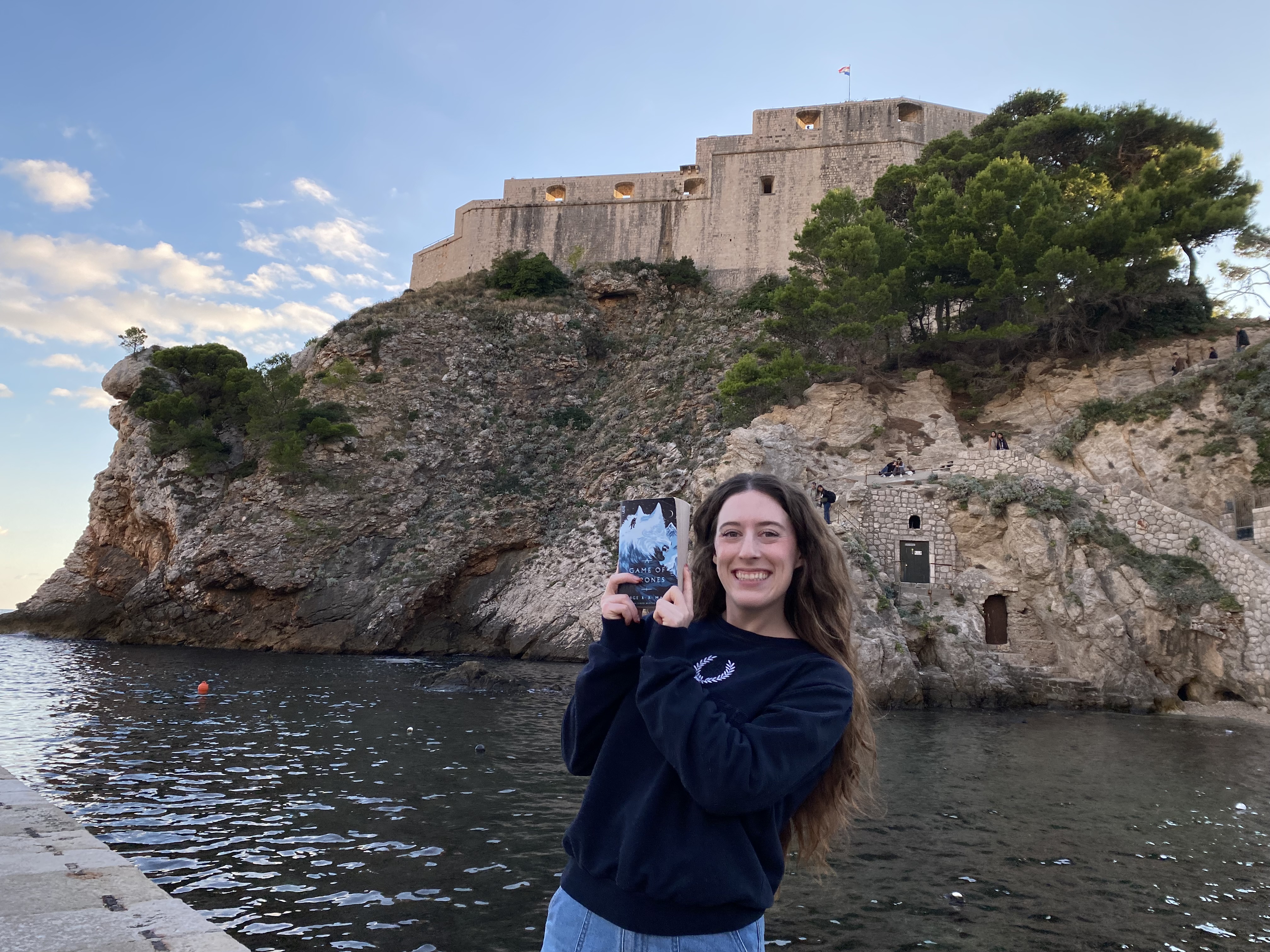 A young woman with brunette hair and a navy sweatshirt holding the Game of Thrones book in front of Fort Lovrijenac – one of the Game of Thrones filming locations in Dubrovnik.