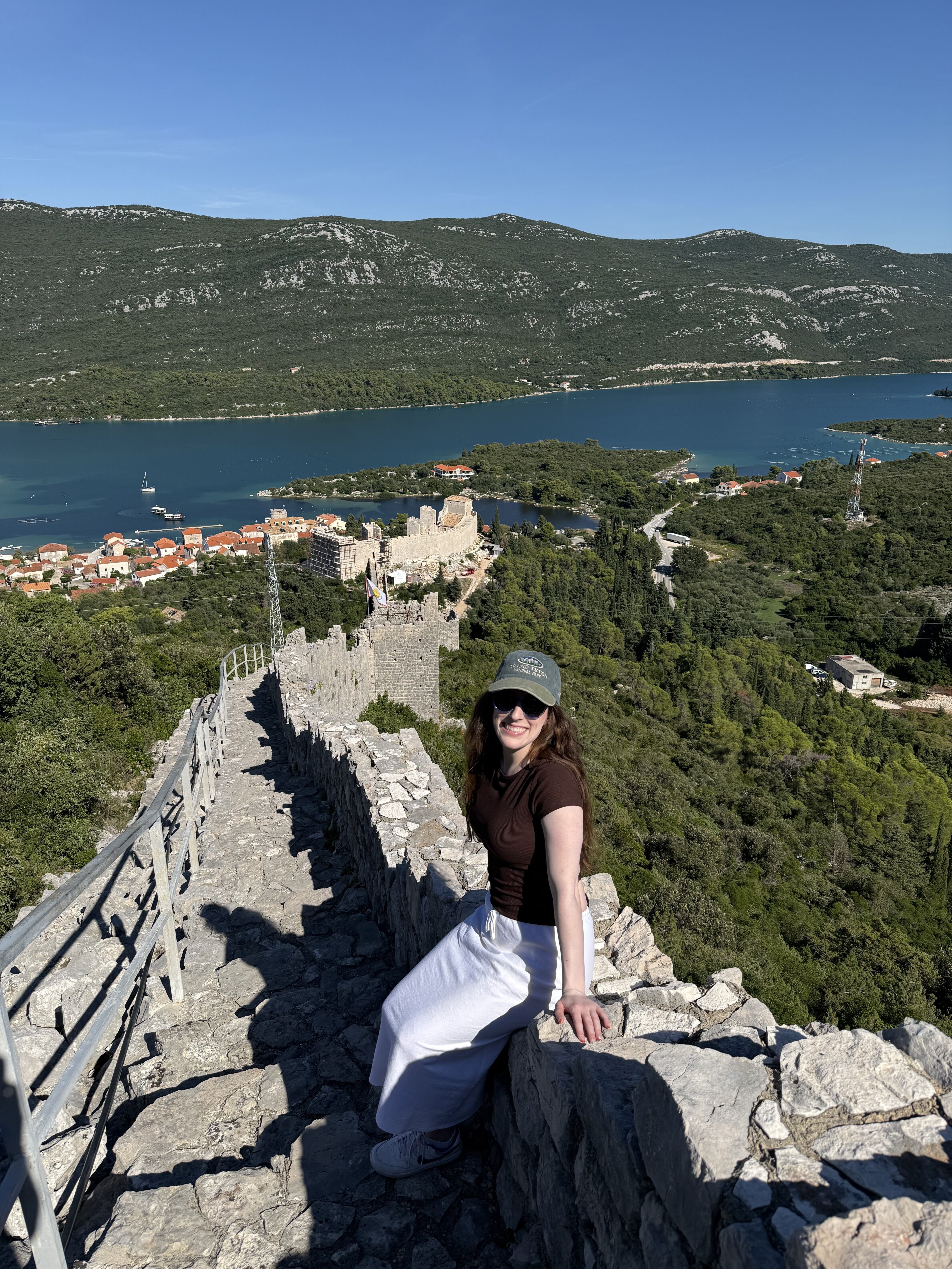A young woman wearing a ball cap, sunglasses, a brown shirt and a white skirt smiling as she sits on the Ston Wall, with Mali Ston in the background.