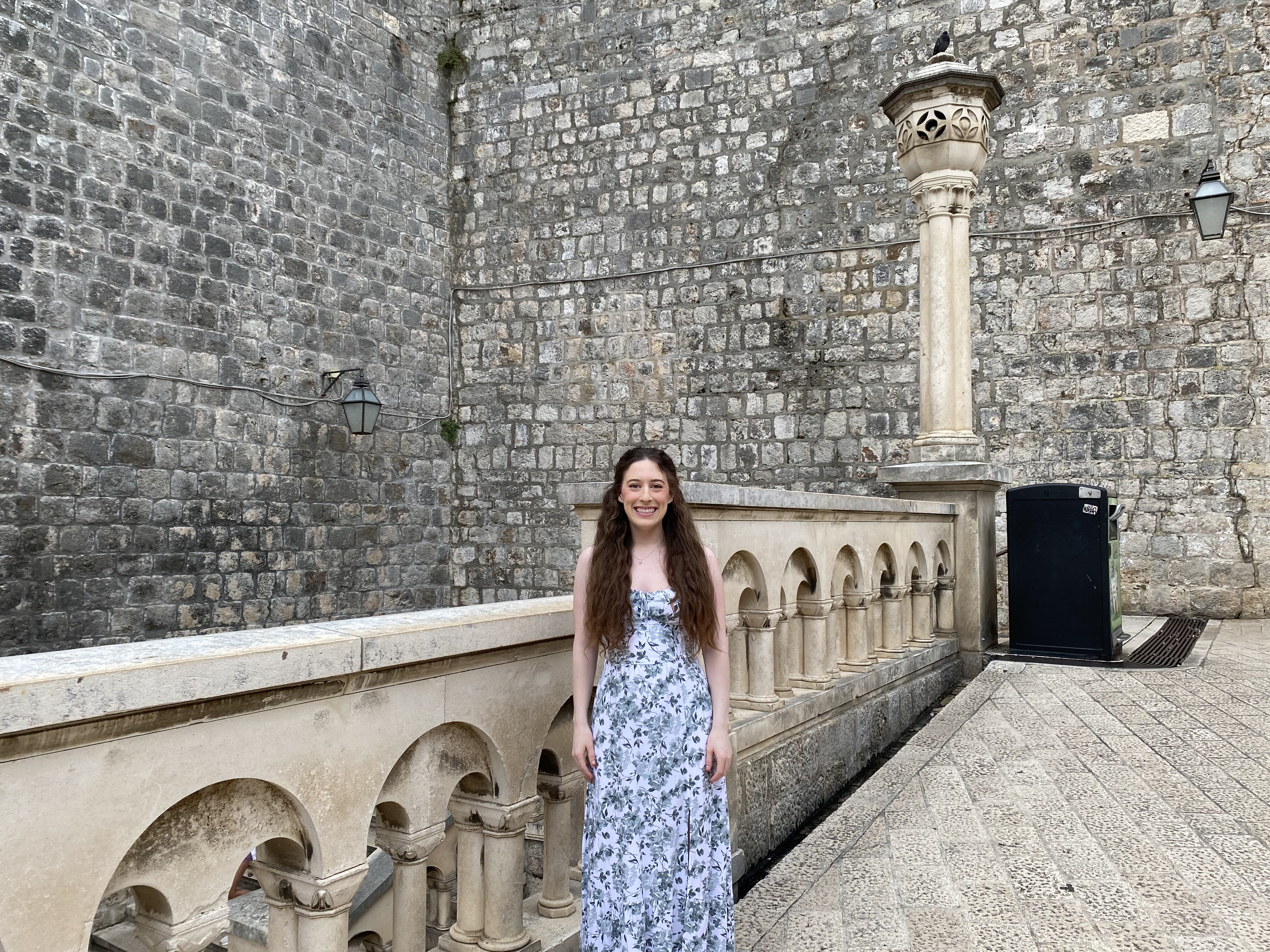 A young woman with brunette hair wearing a white dress with green flowers smiles in front of the stone rail in the Pile Gate entrance.