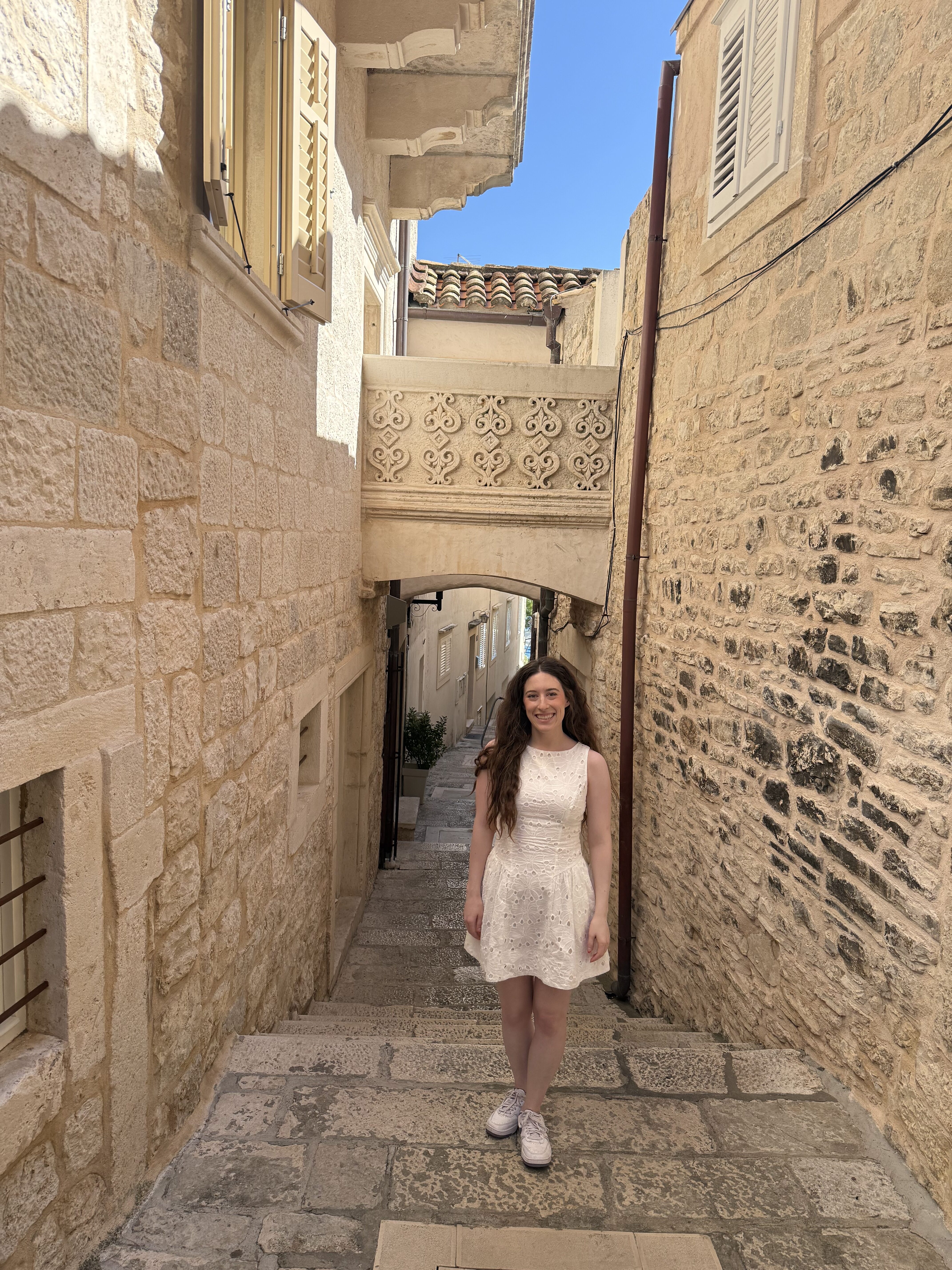 A young brunette woman wearing a white dress smiling on the tan stone streets in Korčula Old Town.