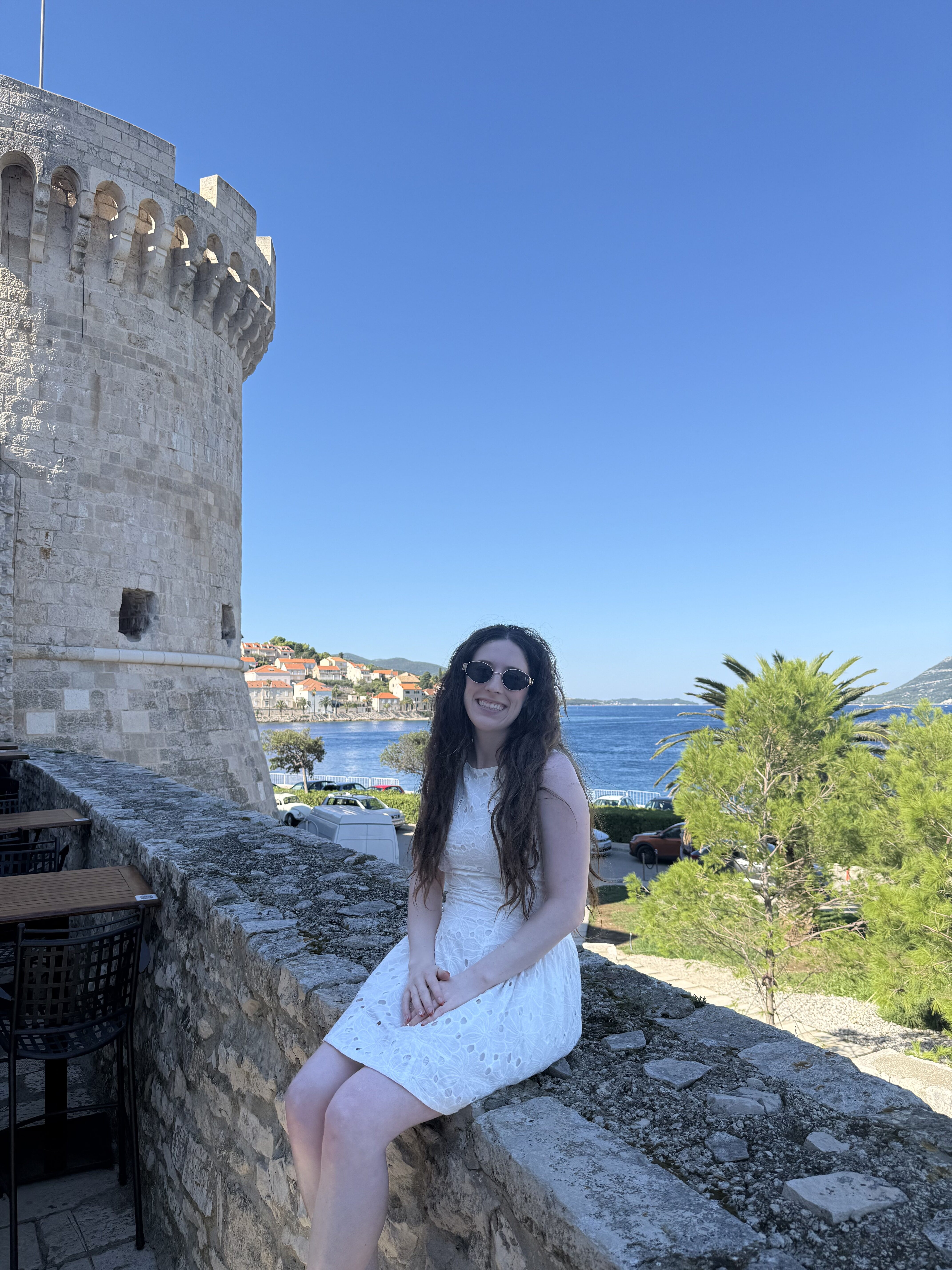 A young woman with brunette hair wearing a short white dress and black sunglasses smiling atop a stone wall in Korčula Old Town.