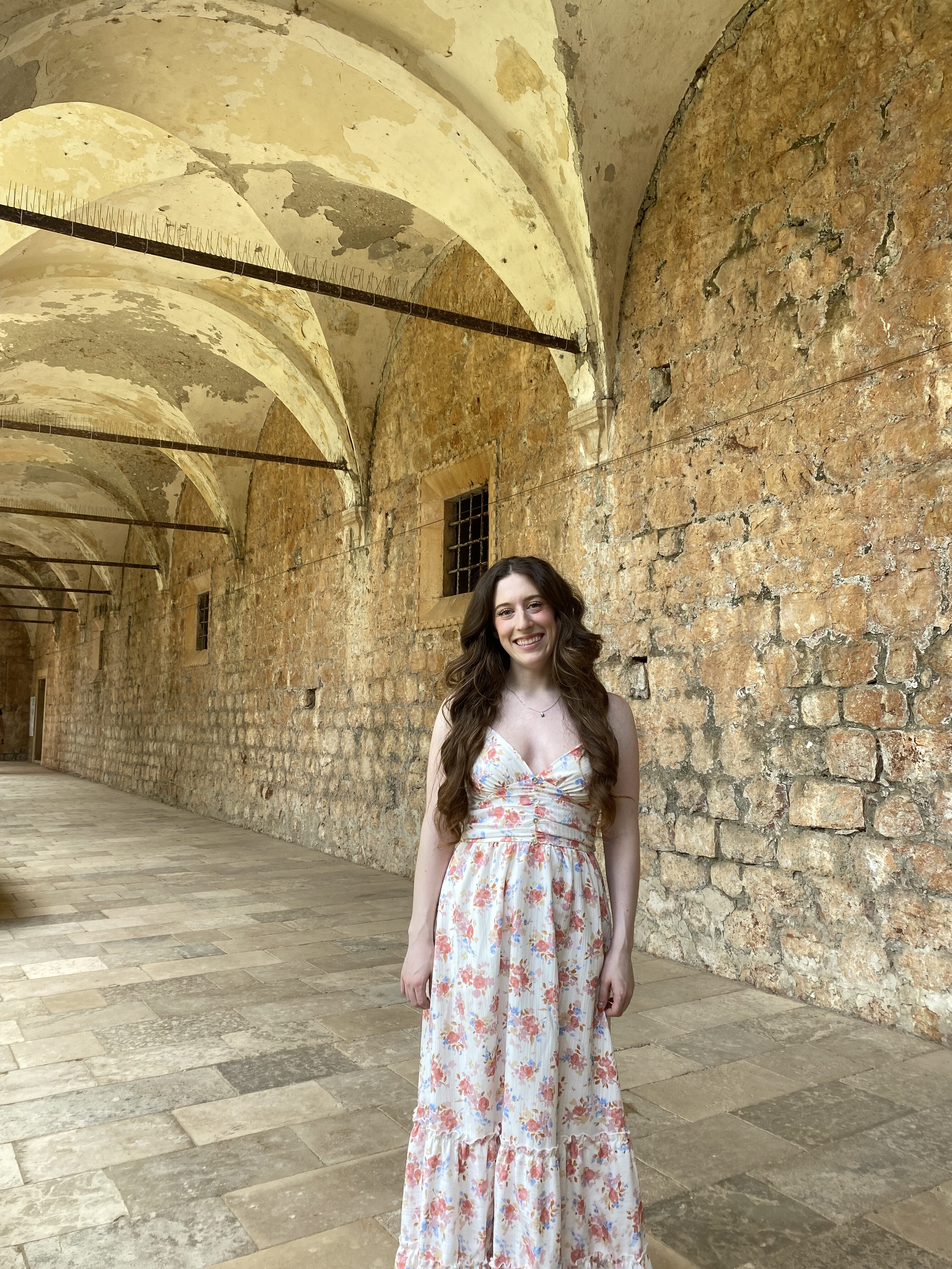 A young woman with brunette hair wearing a white flower dress smiling in the stone portico of the Benedictine Monastery on Lokrum Island.