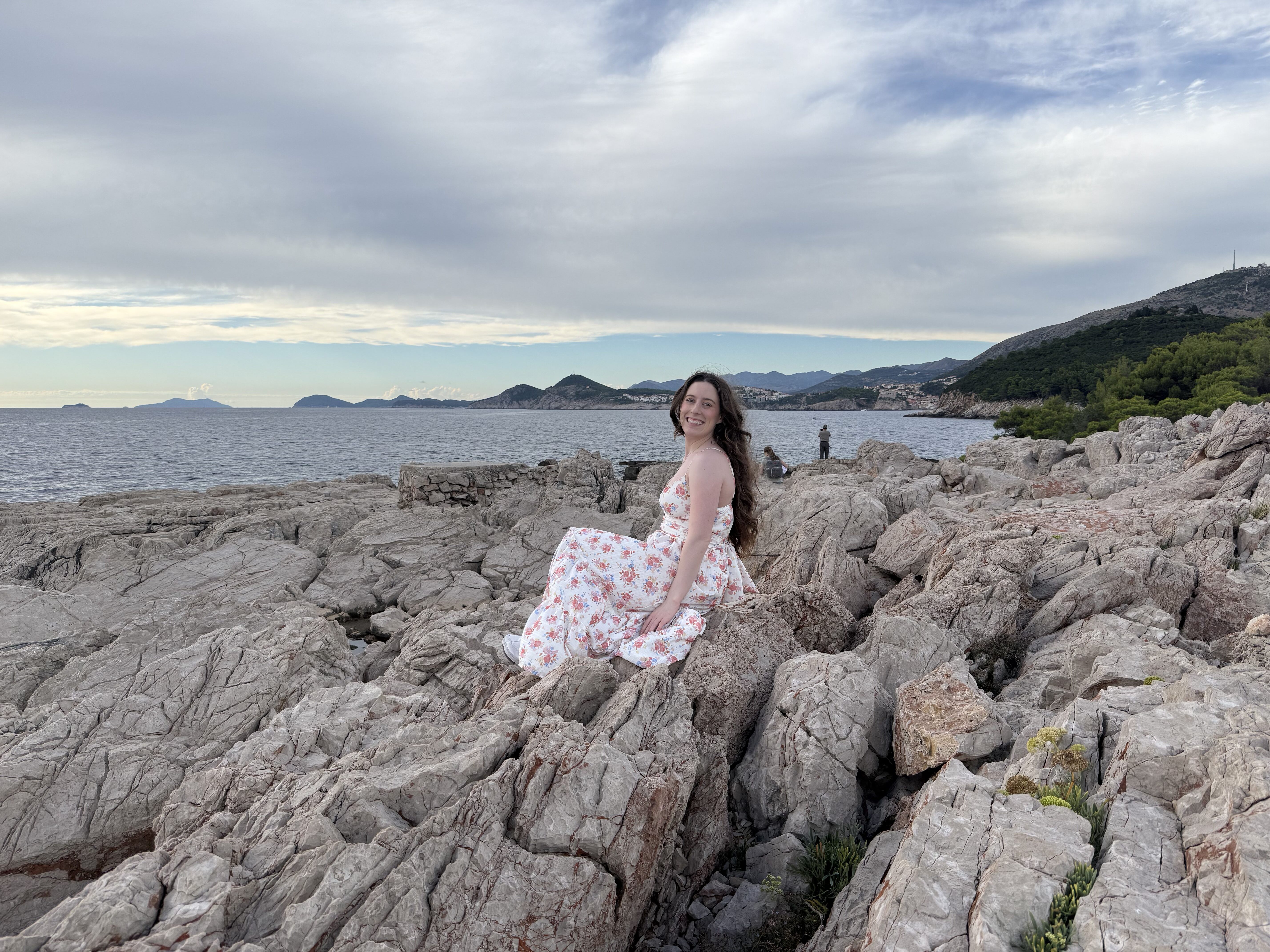 A young woman wearing a white flower dress smiling as she sits upon the rocky coastline of Lokrum Island. Dubrovnik Old Town is in the distance.