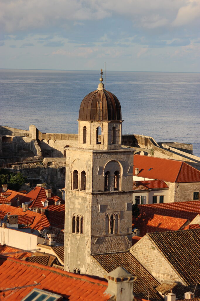 A grey stone bell tower in Dubrovnik with a dark grey dome roof, surrounded by copper-colored roofs in Dubrovnik Old Town with the Adriatic Sea and a blue sky in the background.