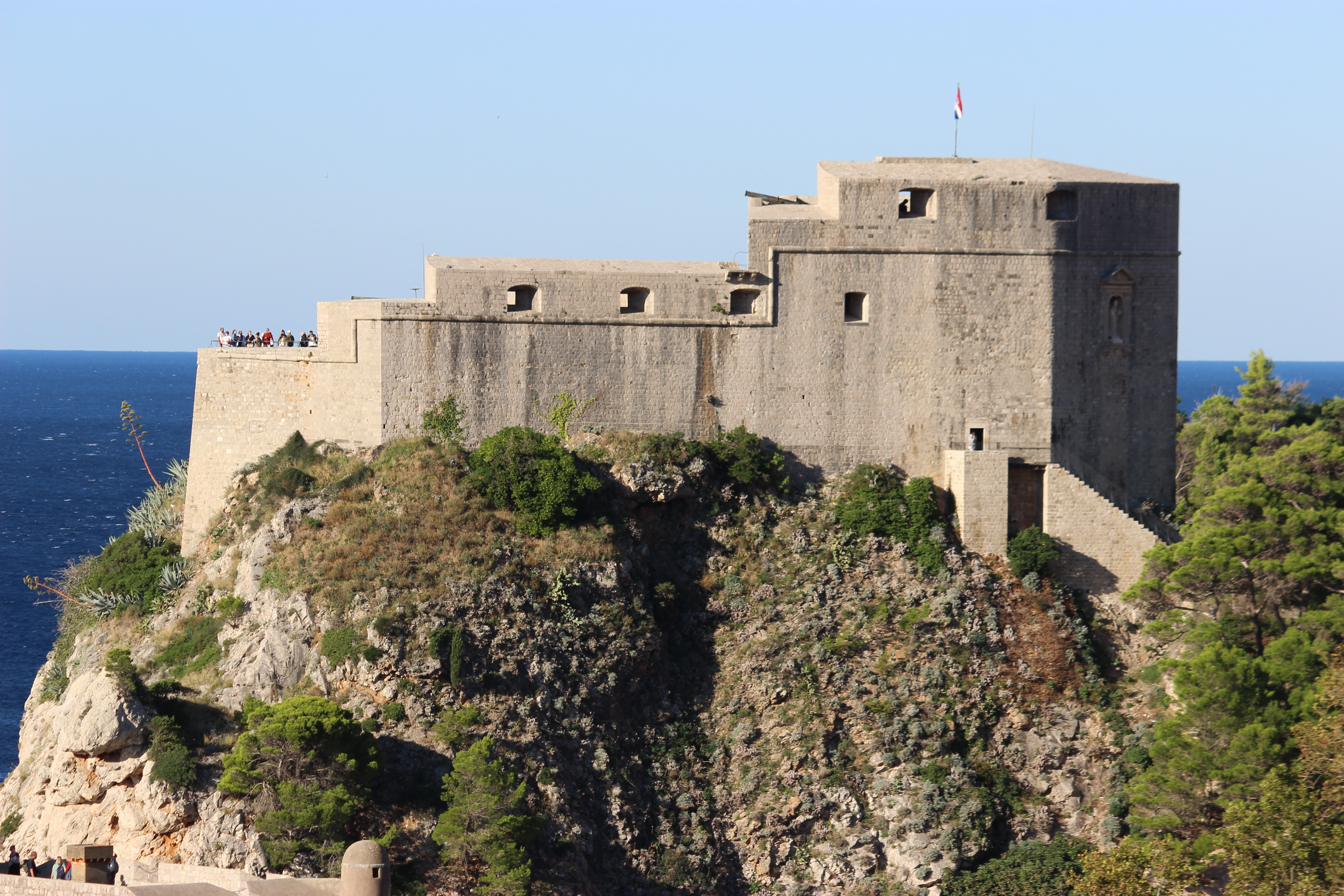 The grey stone Fort Lovrijenac surrounded by green shrubs with the dark blue Adriatic Sea in the background.