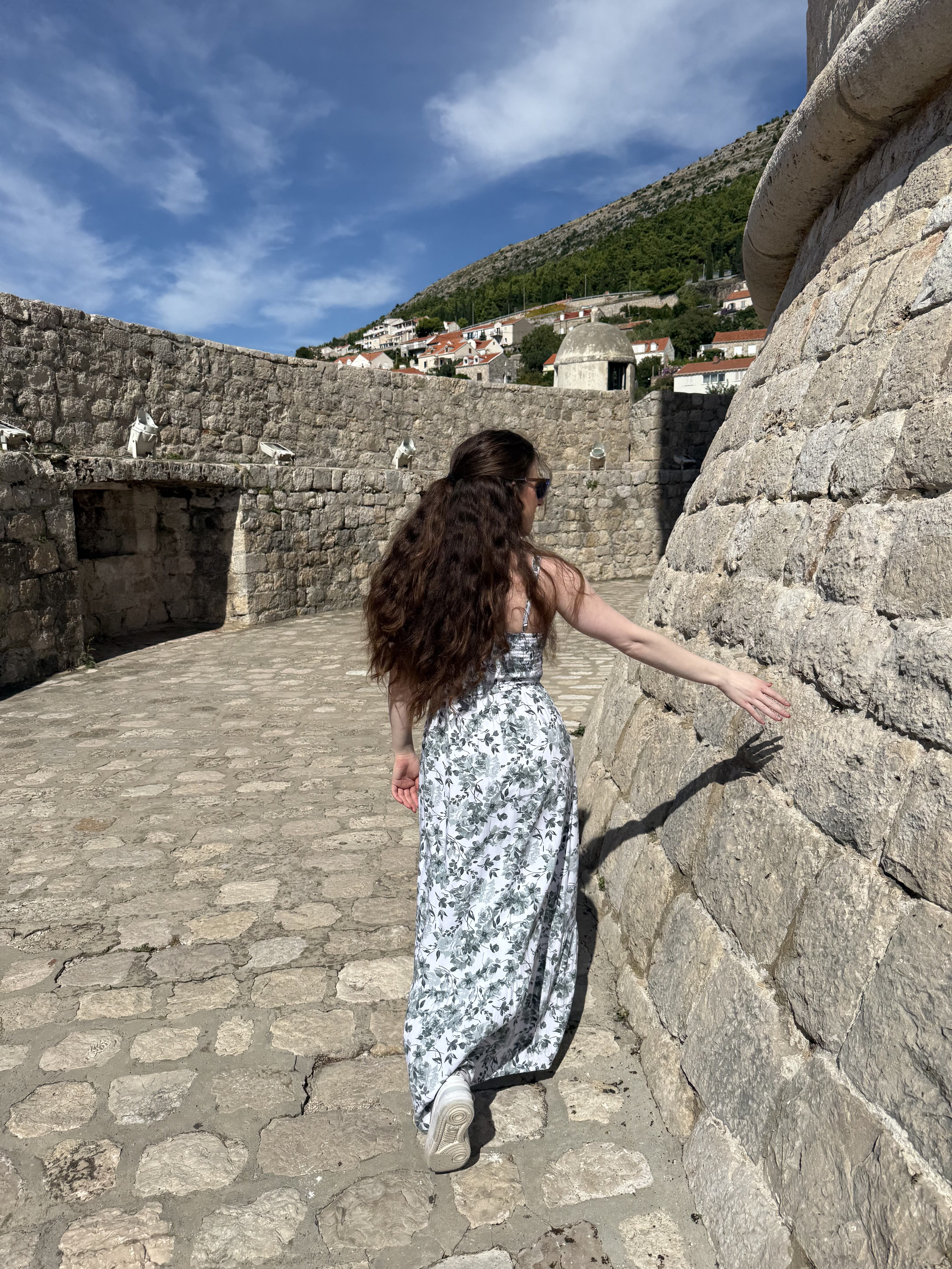A young woman with thick brunette hair walking with her hand outstretched towards the sloped stone wall of the Minčeta Tower.