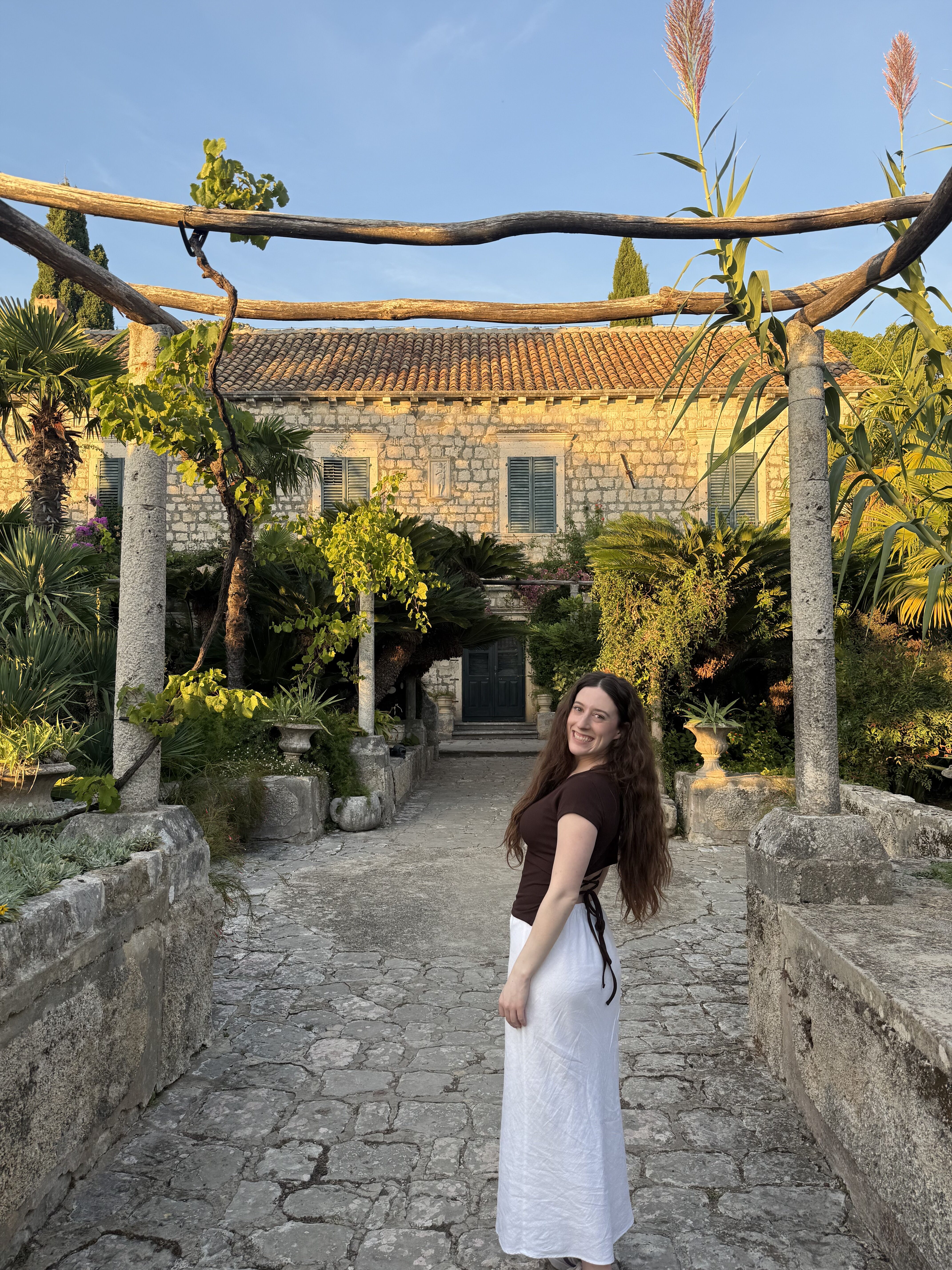 A young woman with thick brunette hair wearing a brown shirt and a white skirt stands under an open canopy in Trsteno Arboretum.
