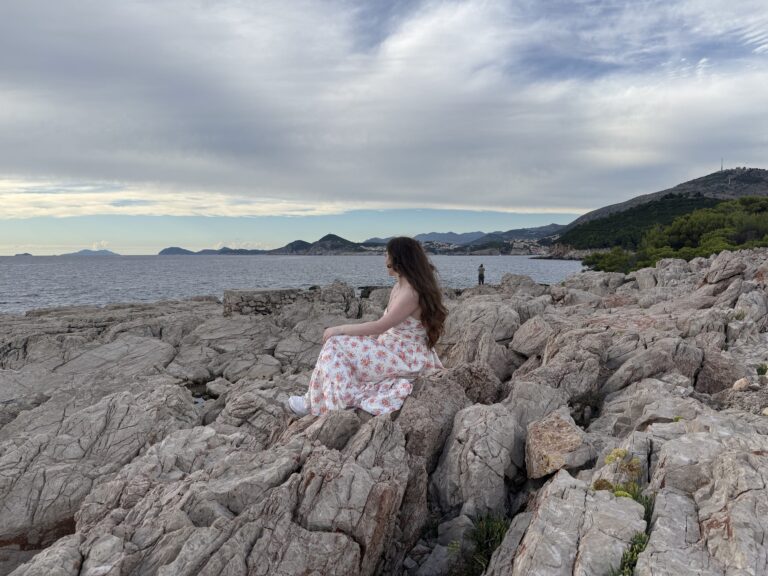 A young woman with thick brunette hair staring out at Dubrovnik Old Town from the rocky shoreline of Lokrum Island.