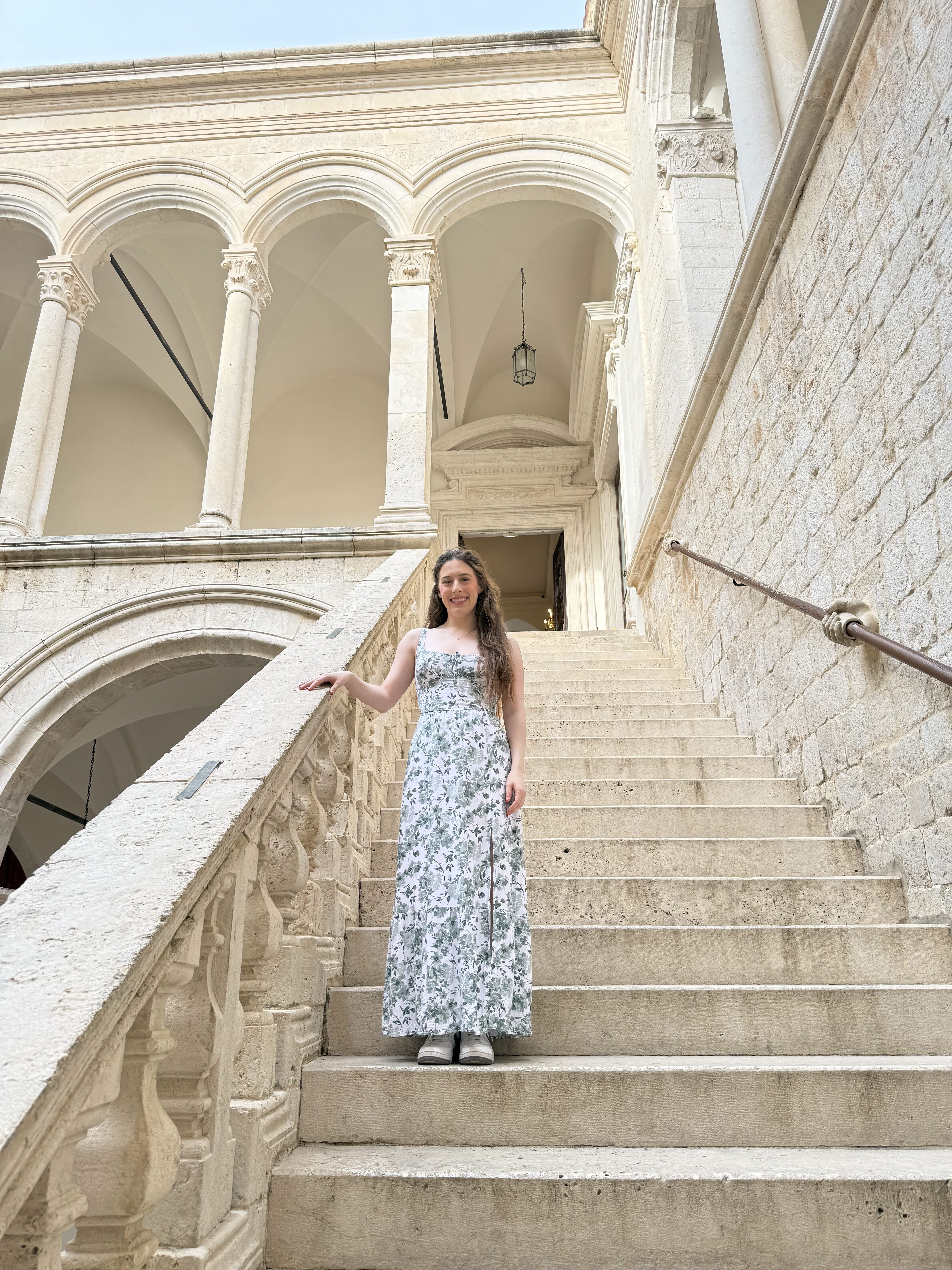 A young woman wearing a white dress with green flowers smiling on an off-white stone staircase in Rector's Palace.