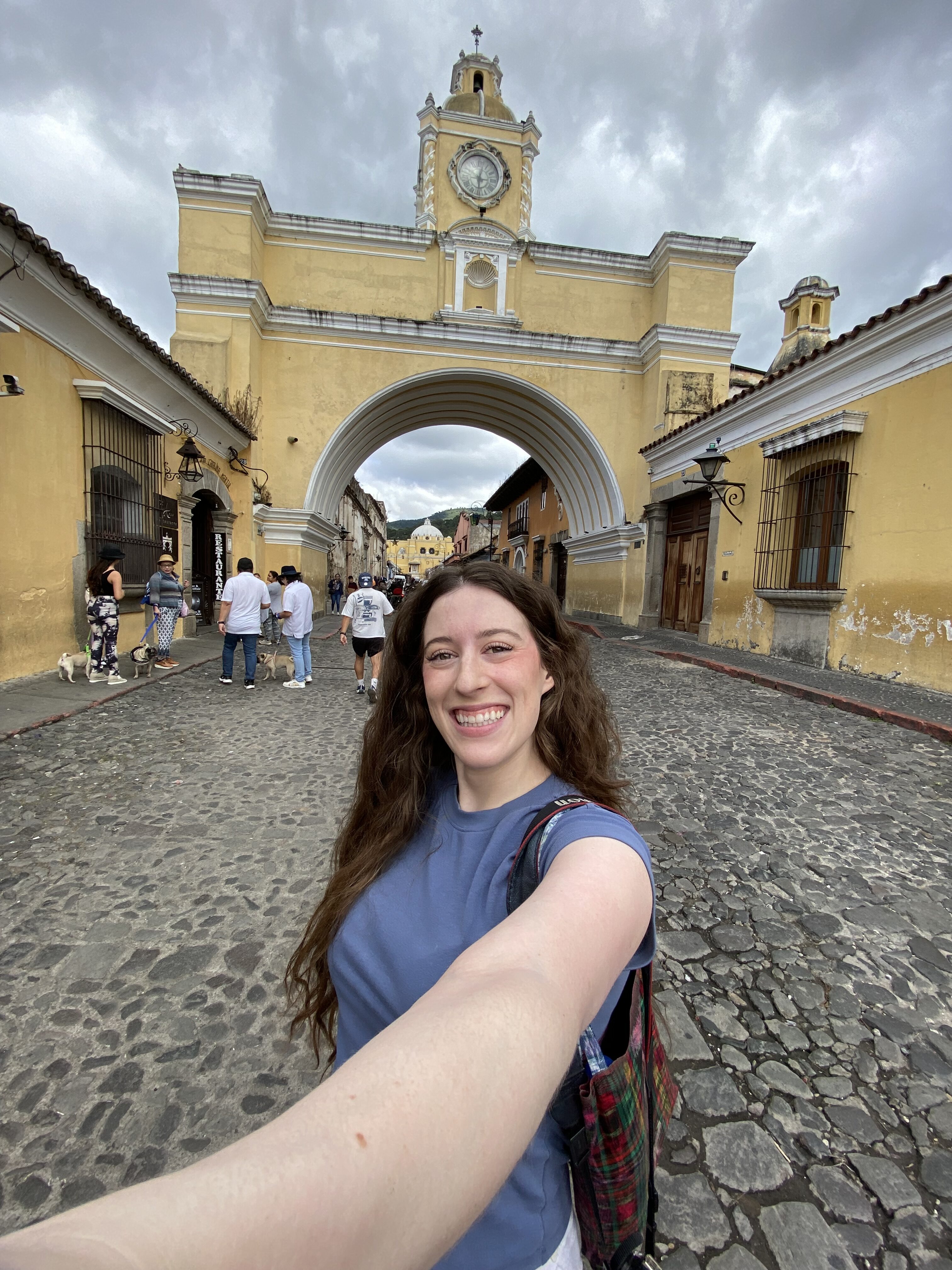 A young brunette woman smiling in a selfie in front of the yellow Arco de Santa Catalina.