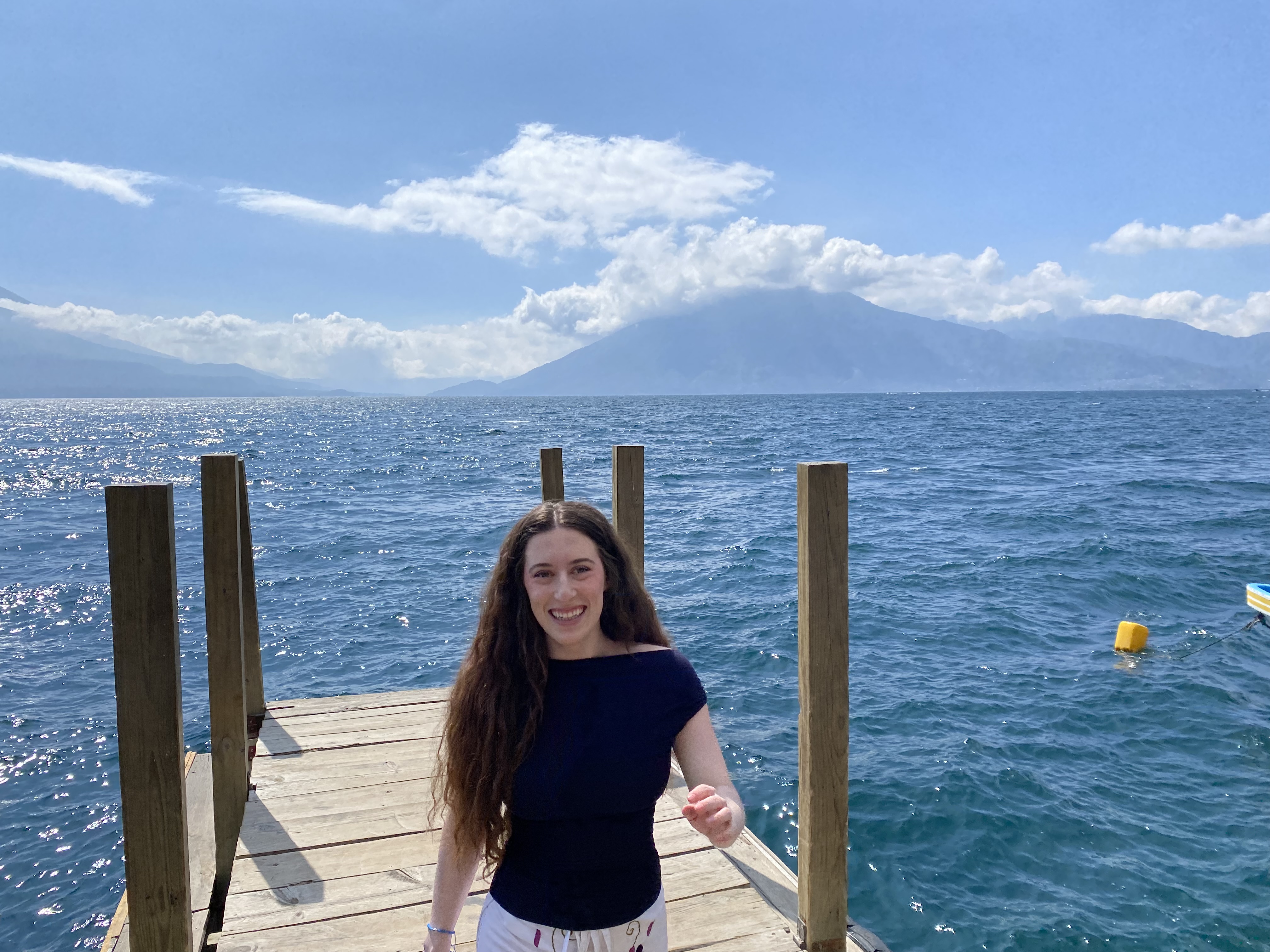 Bella, smiling and walking towards the camera on the dock of La Casa del Mundo with Lake Atitlán in the background.