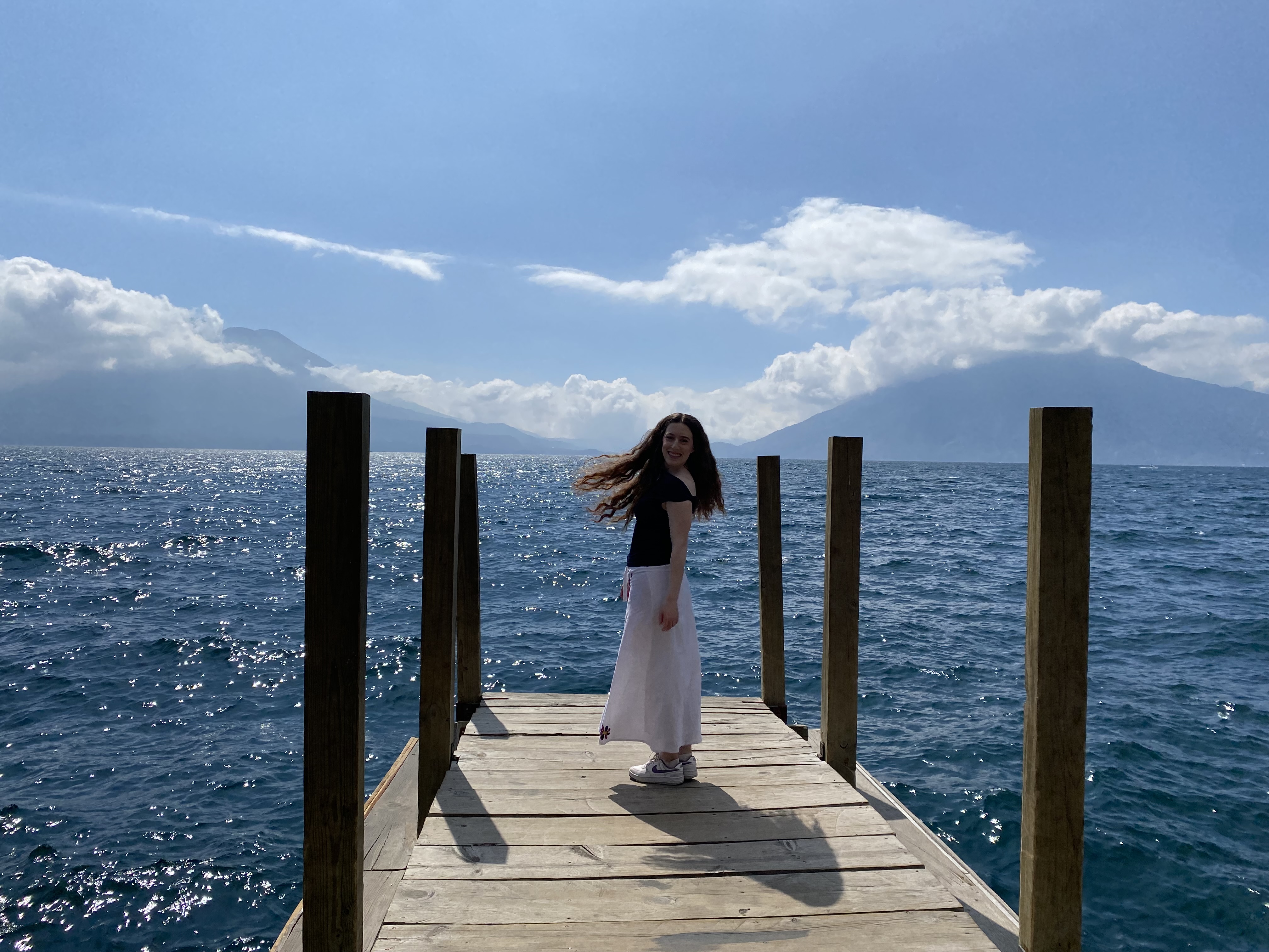 A young woman wearing a white skirt and a blue shirt standing on the dock in front of Lake Atitlán, swishing her hair with two large volcanoes in the background.