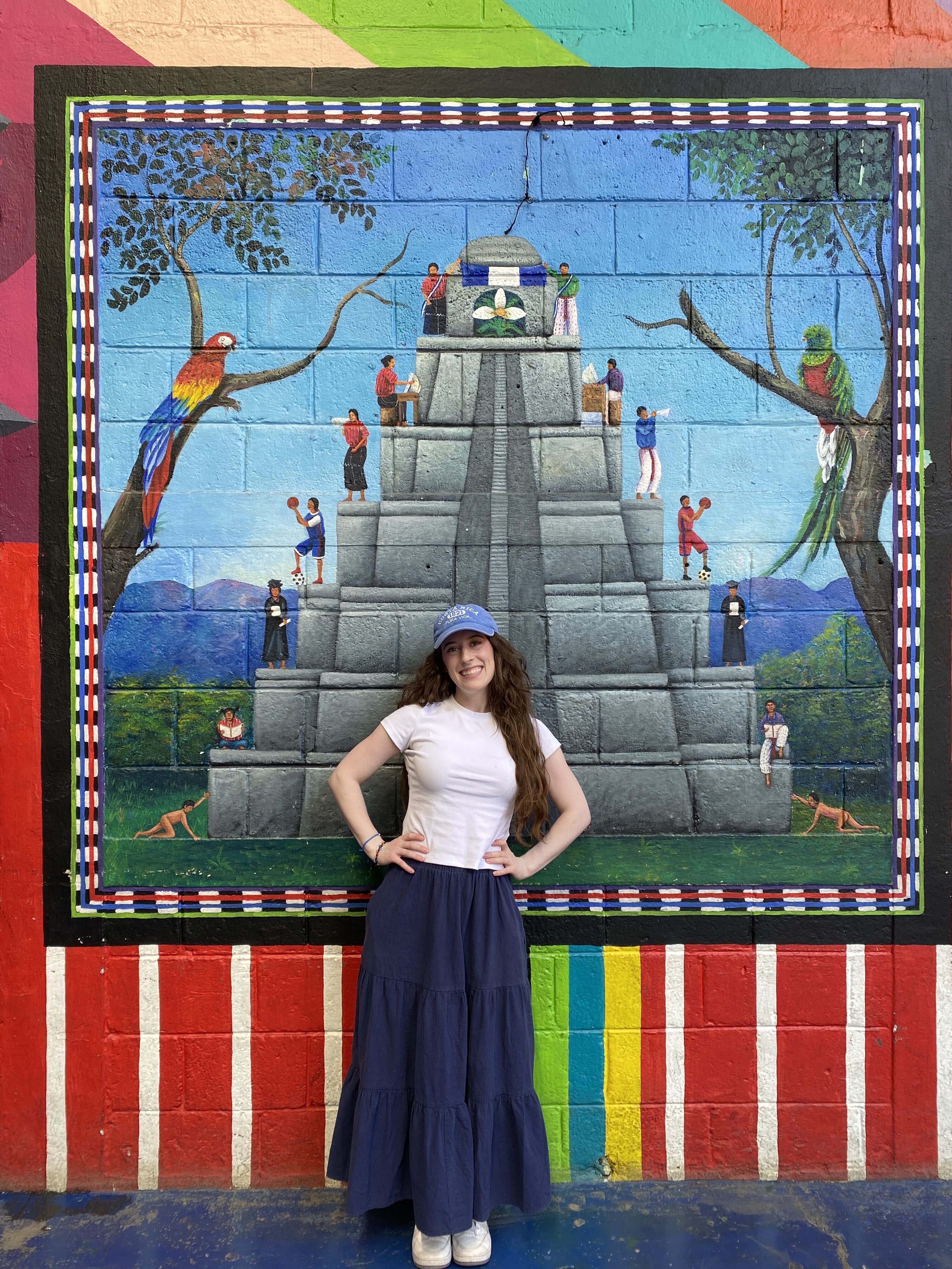 A young woman wearing a white shirt, blue skirt and a hat with long brunette hair smiling in front of a colorful Tikal mural in San Juan (Lake Atitlán).