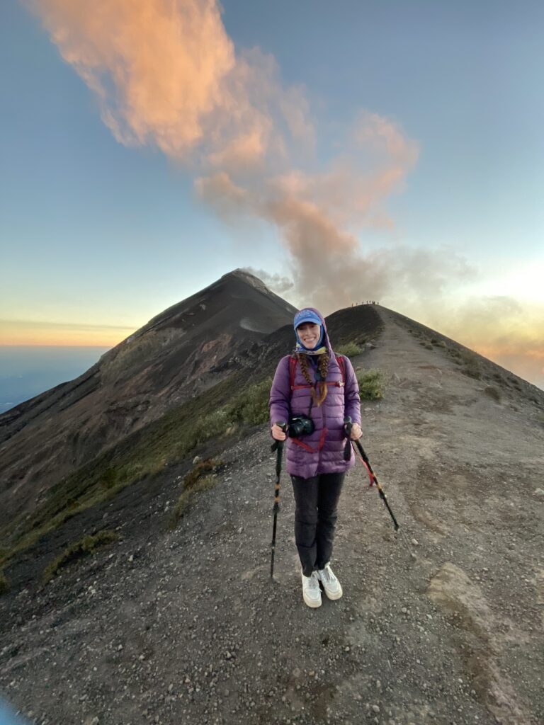 A young woman wearing a purple coat and black pants holds her hiking poles as she smiles in front of Fuego volcano at sunset.