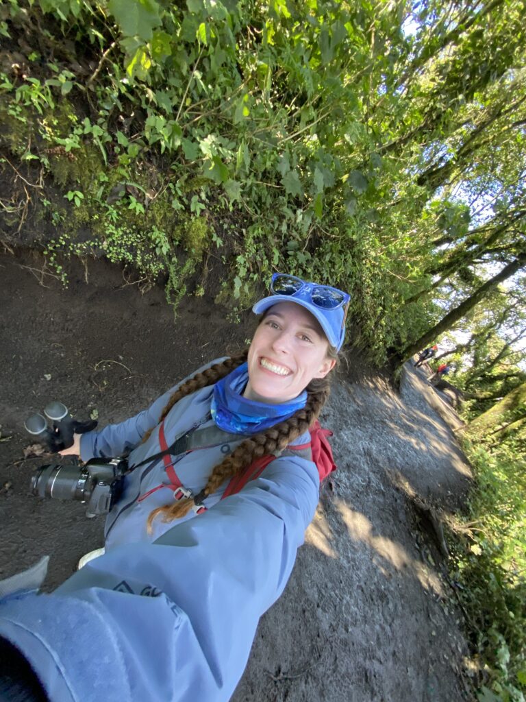 A young woman with two braids wearing a blue raincoat and a blue hat smiling amidst the tree-covered Acatenango hiking trail.