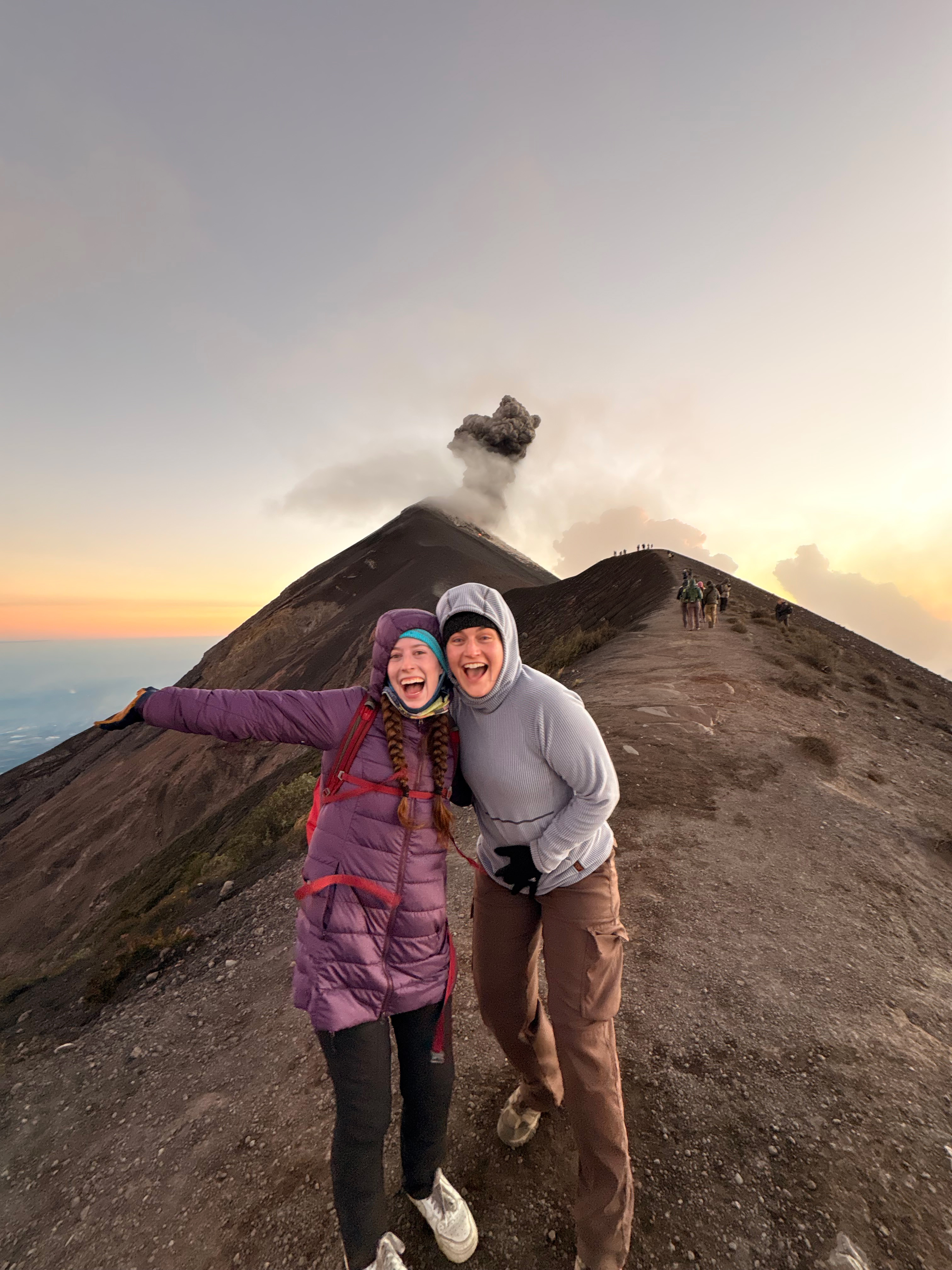 A pair of ecstatic hikers smiling on the ridge of Fuego Volcano while smoke plumes from the top.