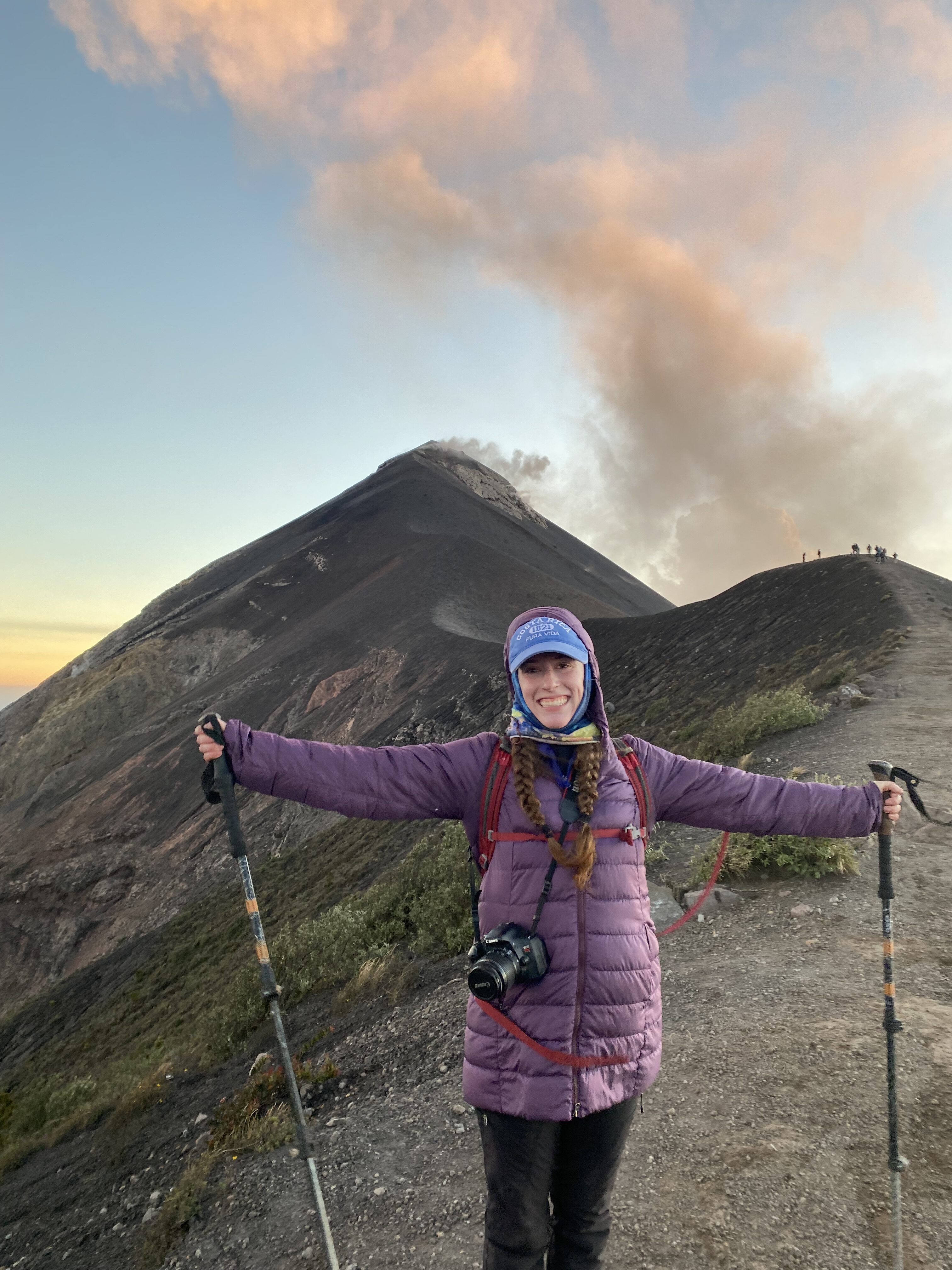 A young woman with a camera slung around her neck wearing a purple coat holding hiking poles in front of smoking Fuego Volcano in Guatemala.