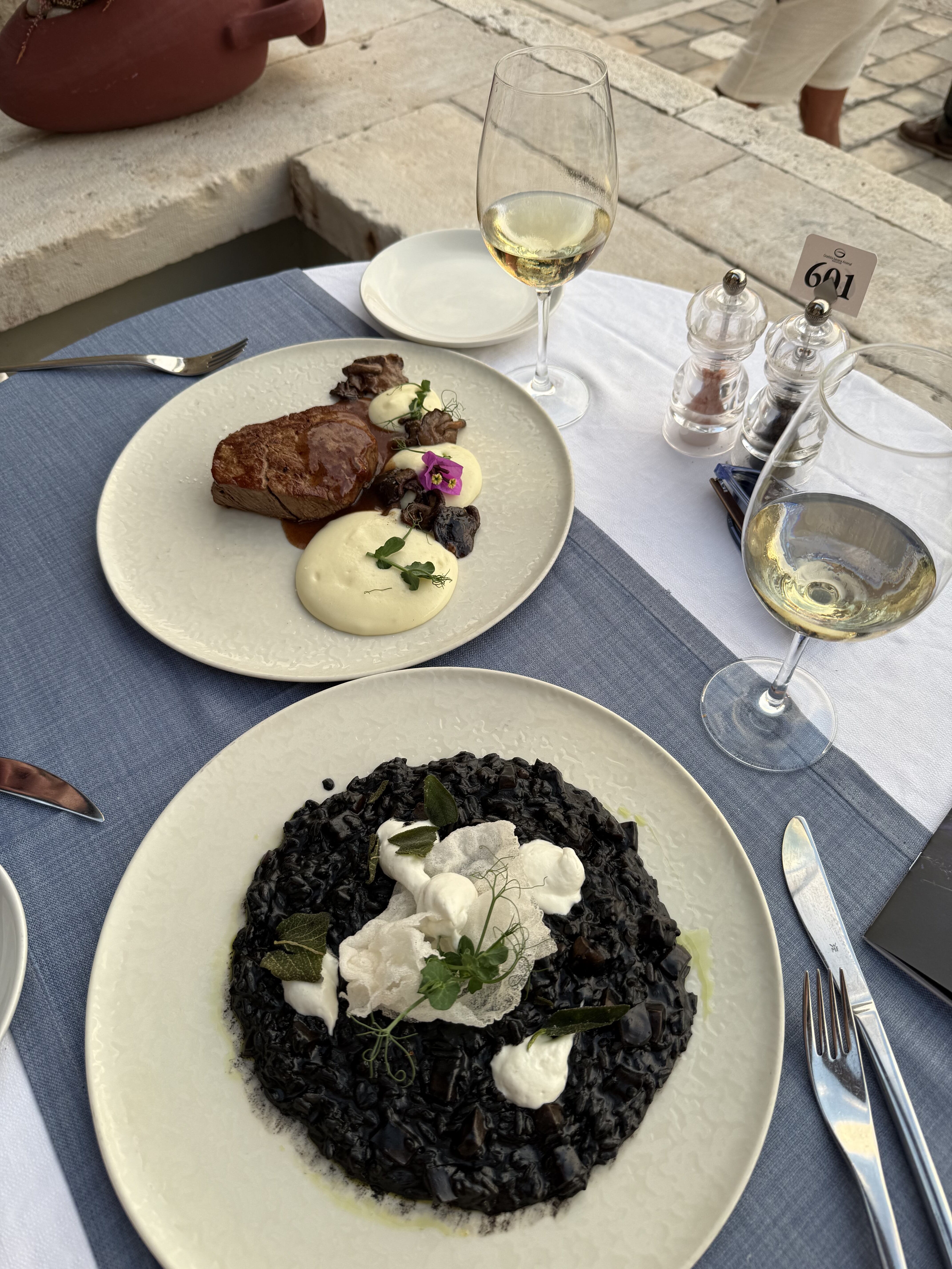 Black cuttlefish risotto and filet mignon plated on a blue tablecloth with glasses of Croatian white wine at Gradska Arsenal.