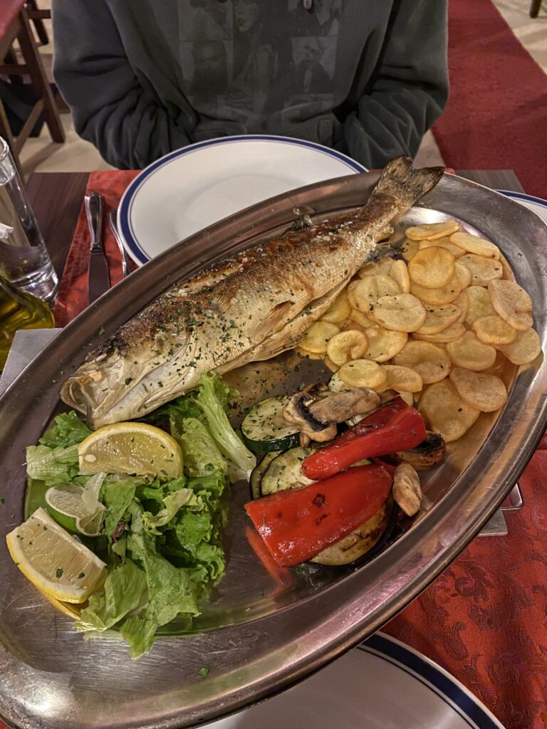 A grilled fish on a platter with potato fries, greens and roasted red peppers at Konoba Moby Dick.