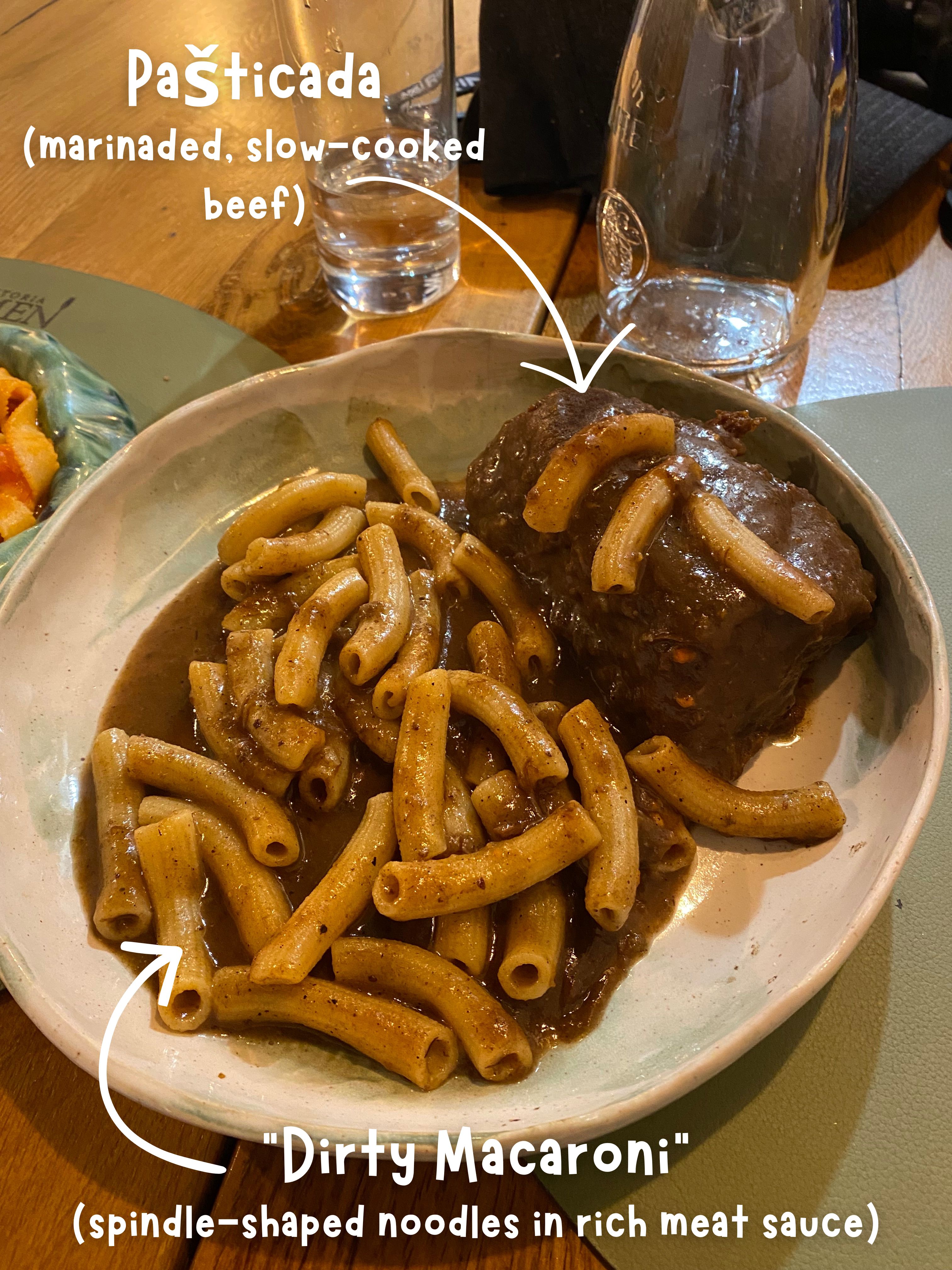 Text and arrows overlayed on an image of a dish containing noodles in rich sauce with slow-roasted beef. The text reads, "Pašticada, marinaded, slow-cooked beef" and "'Dirty Macaroni', spindle-shaped noodles in rich meat sauce".