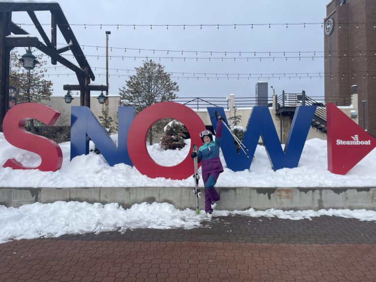 A skier wearing a three-colored ski suit (turquoise, blue and purple) posing in front of a Steamboat "SNOW" sign.