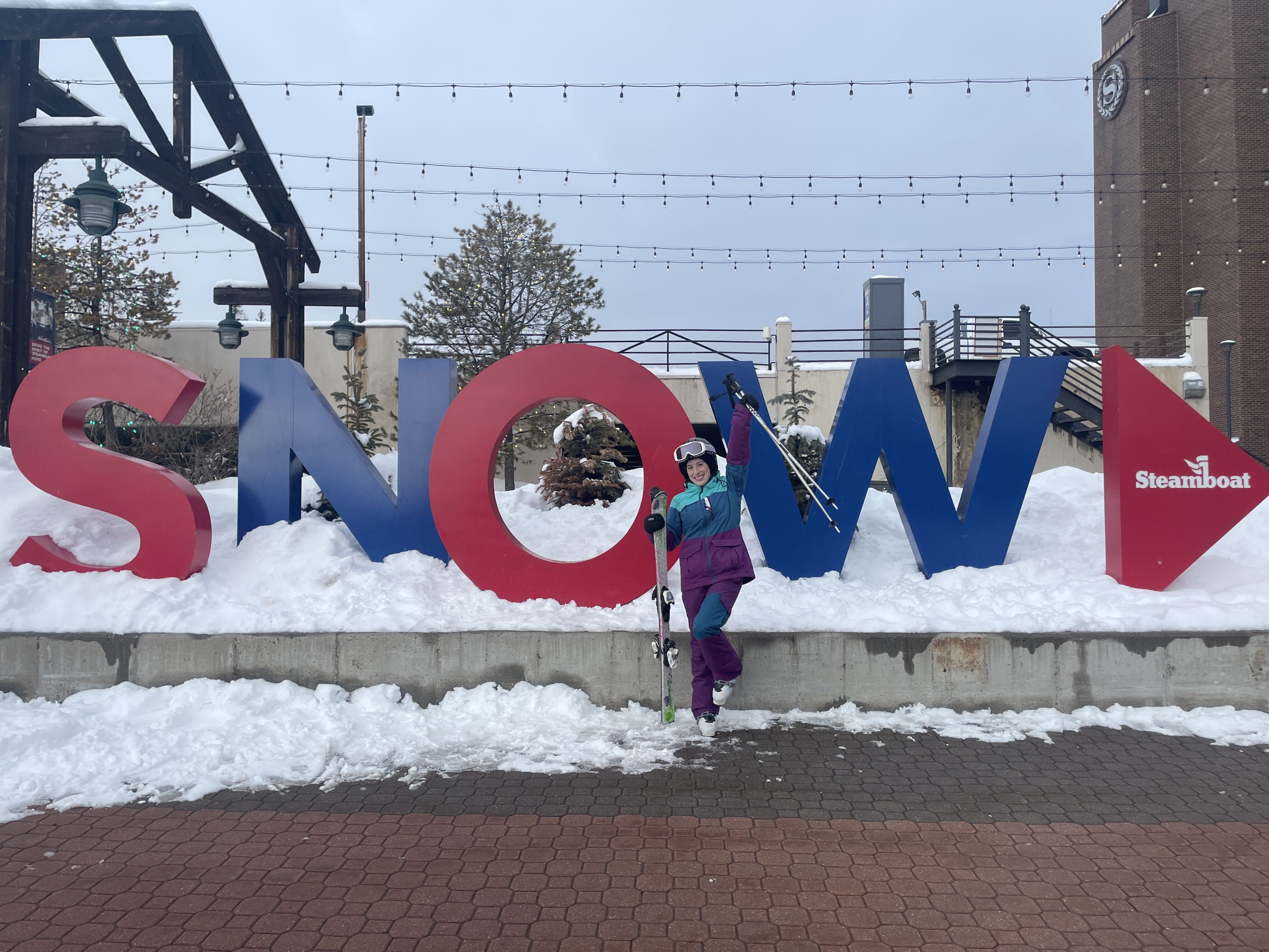 A skier wearing a three-colored ski suit (turquoise, blue and purple) posing in front of a Steamboat "SNOW" sign.