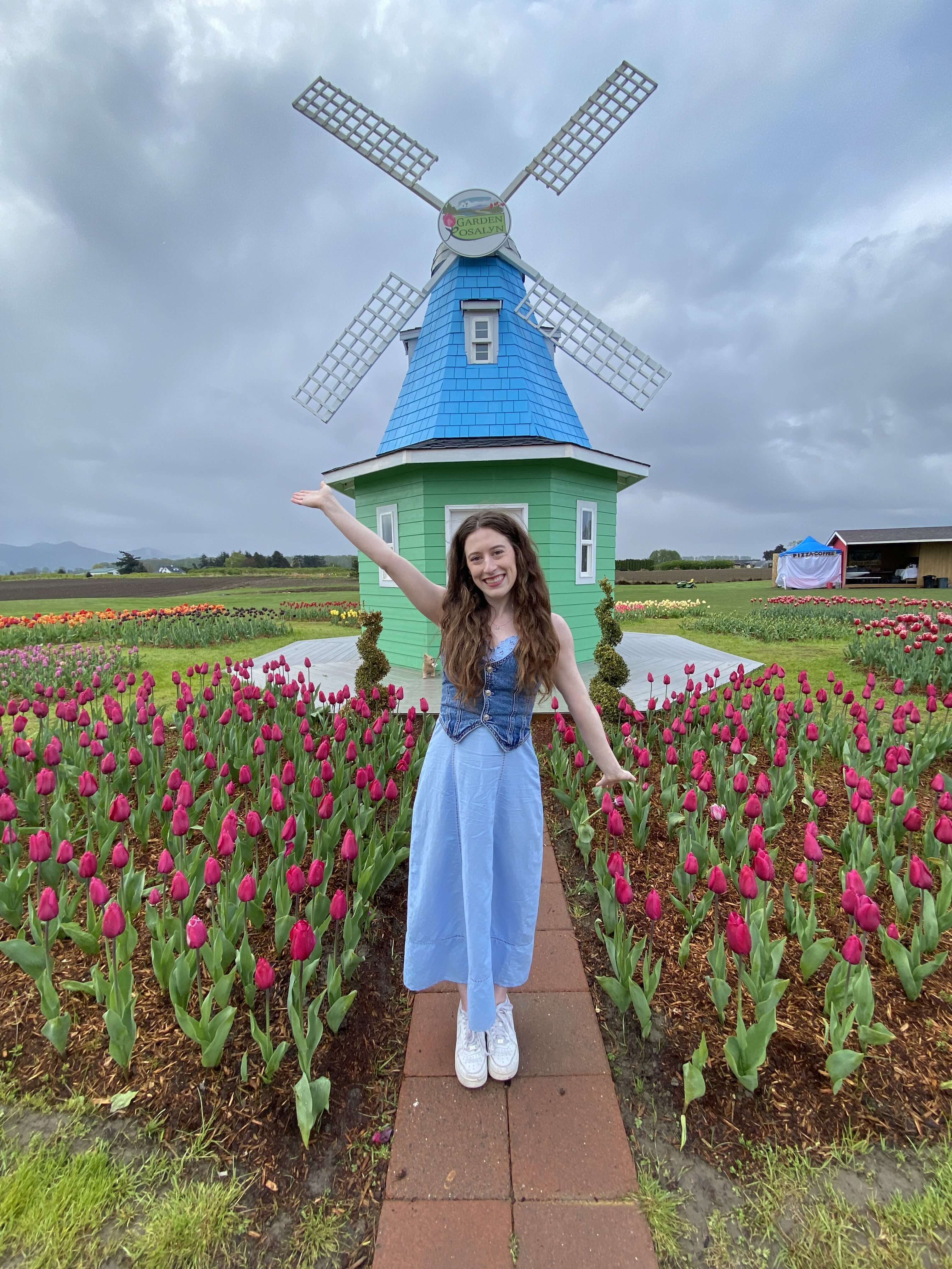 Bella posing in a blue dress with a jean vest in front of Garden Rosalyn's blue/green windmill.