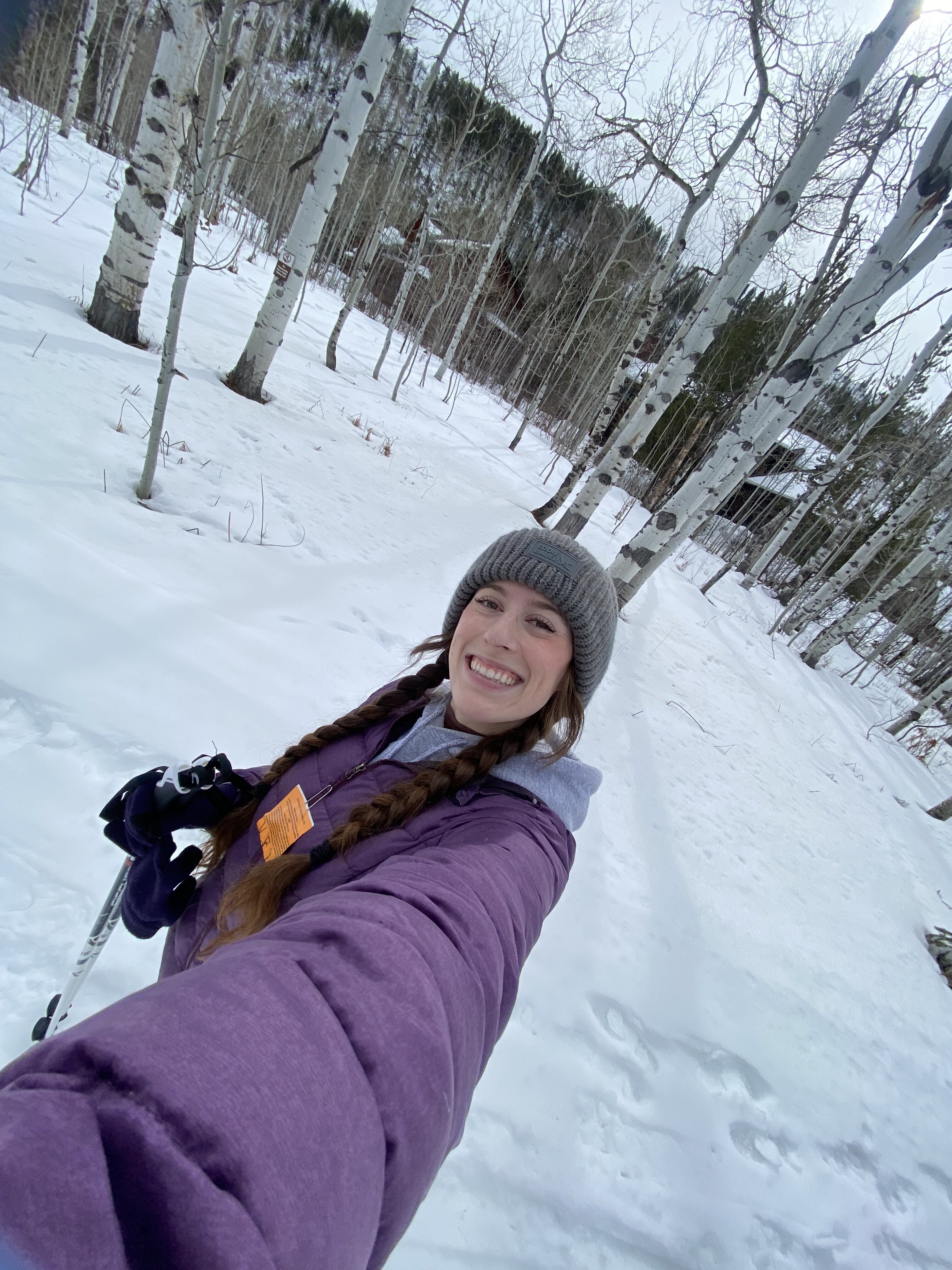 Bella taking a selfie in her purple coat and grey beanie holding Nordic ski poles in front of a winter landscape filled with leaf-less Aspen trees.