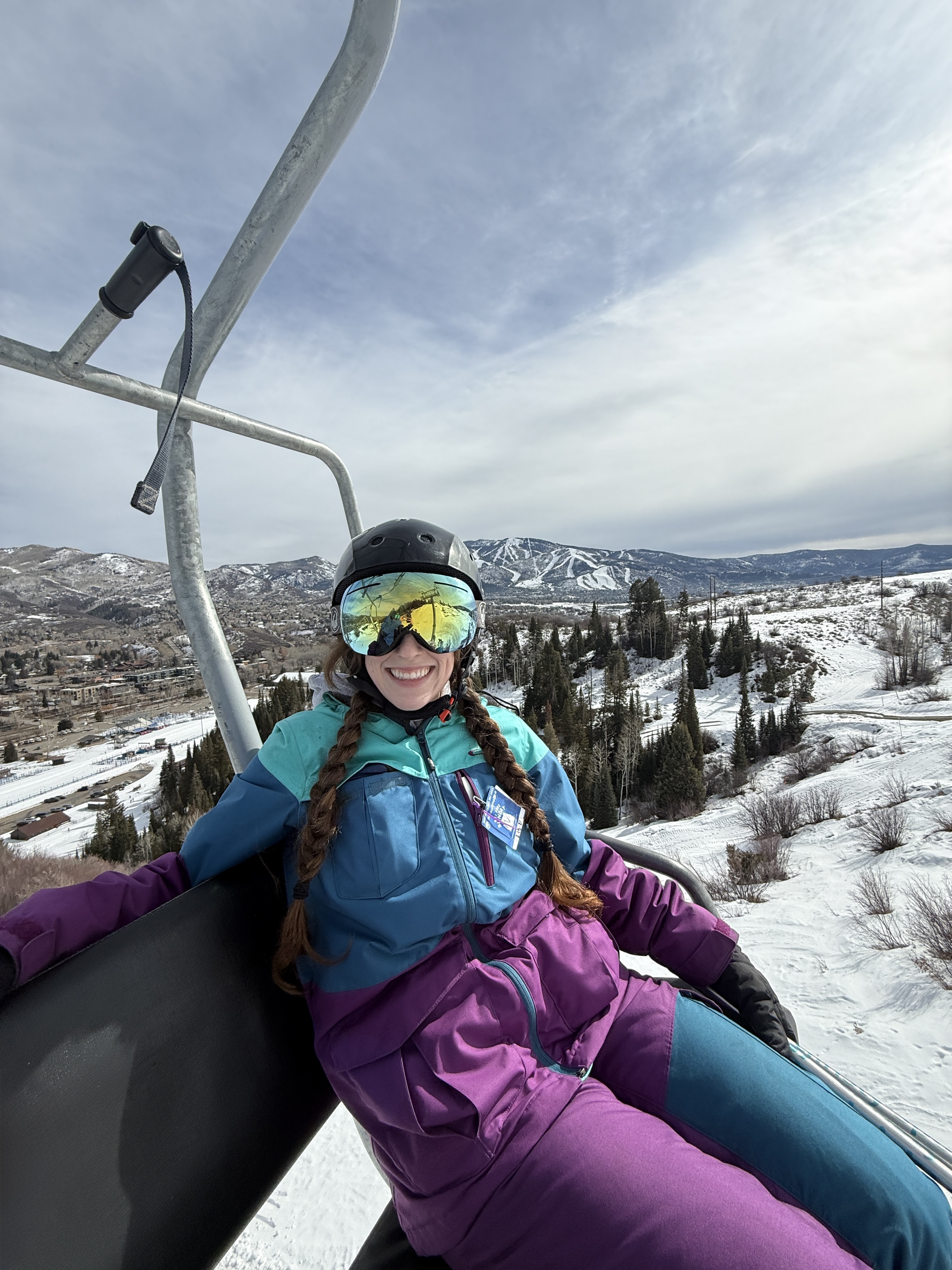 Bella, wearing a three-toned (turqoise, blue and purple) ski outfit with colorful reflective goggles smiling from a lift at Howelsen Hill, Steamboat Springs, Colorado.