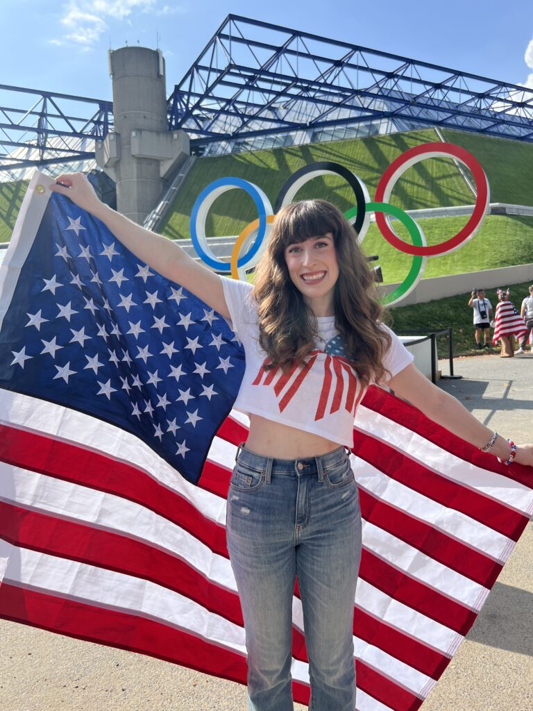 Bella smiling in front of the Olympic rings with an American flag (Paris 2024 Olympics)
