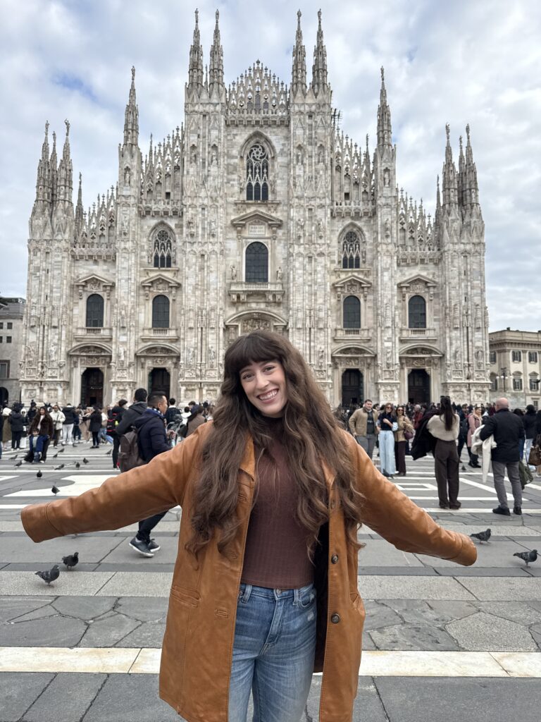 Bella in a brown leather jacket smiling in front of the Milano Duomo.