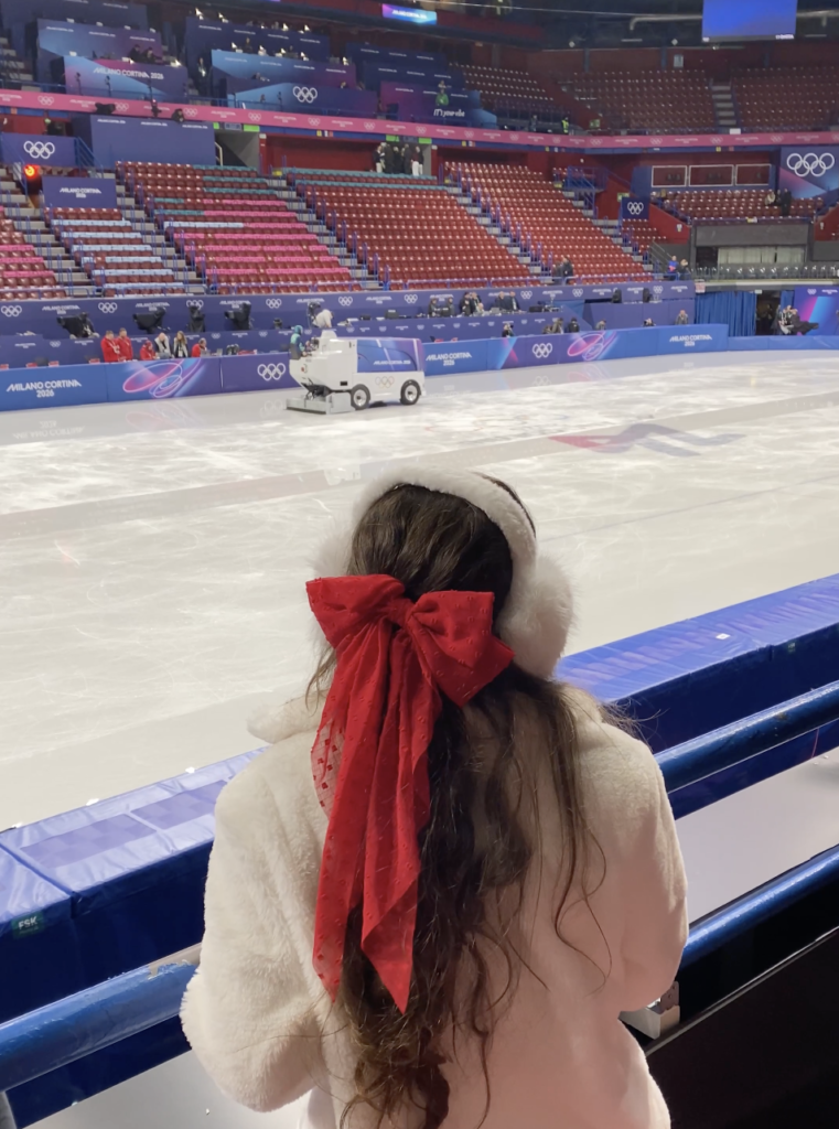 Bella gazing out the the 2026 Milano-Cortina Winter Olympics figure skating arena with a bright red bow and white ear muffs.
